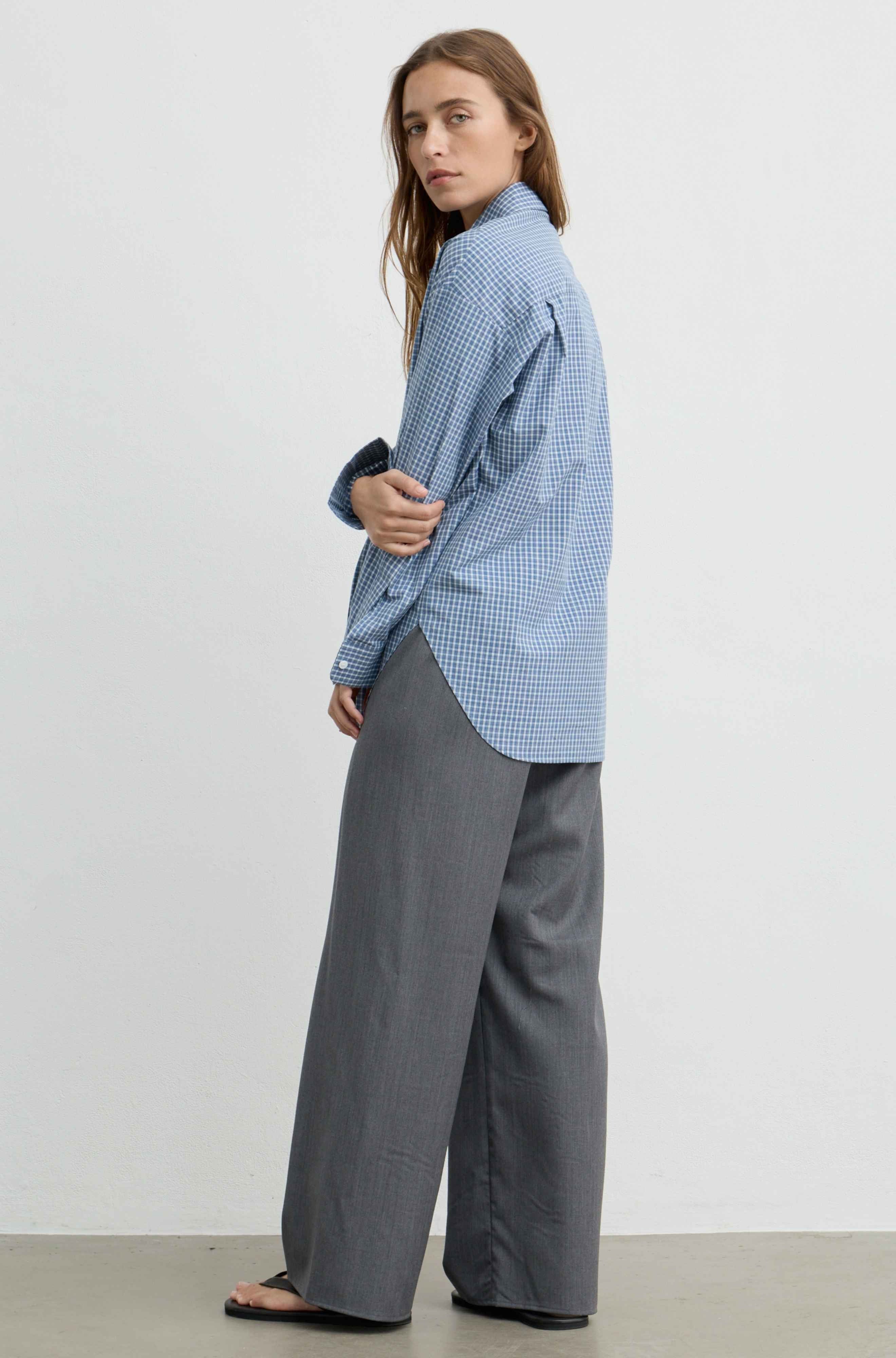 A woman modeling a light blue and white gingham long-sleeve shirt paired with wide-leg gray trousers, standing against a plain light gray studio wall.