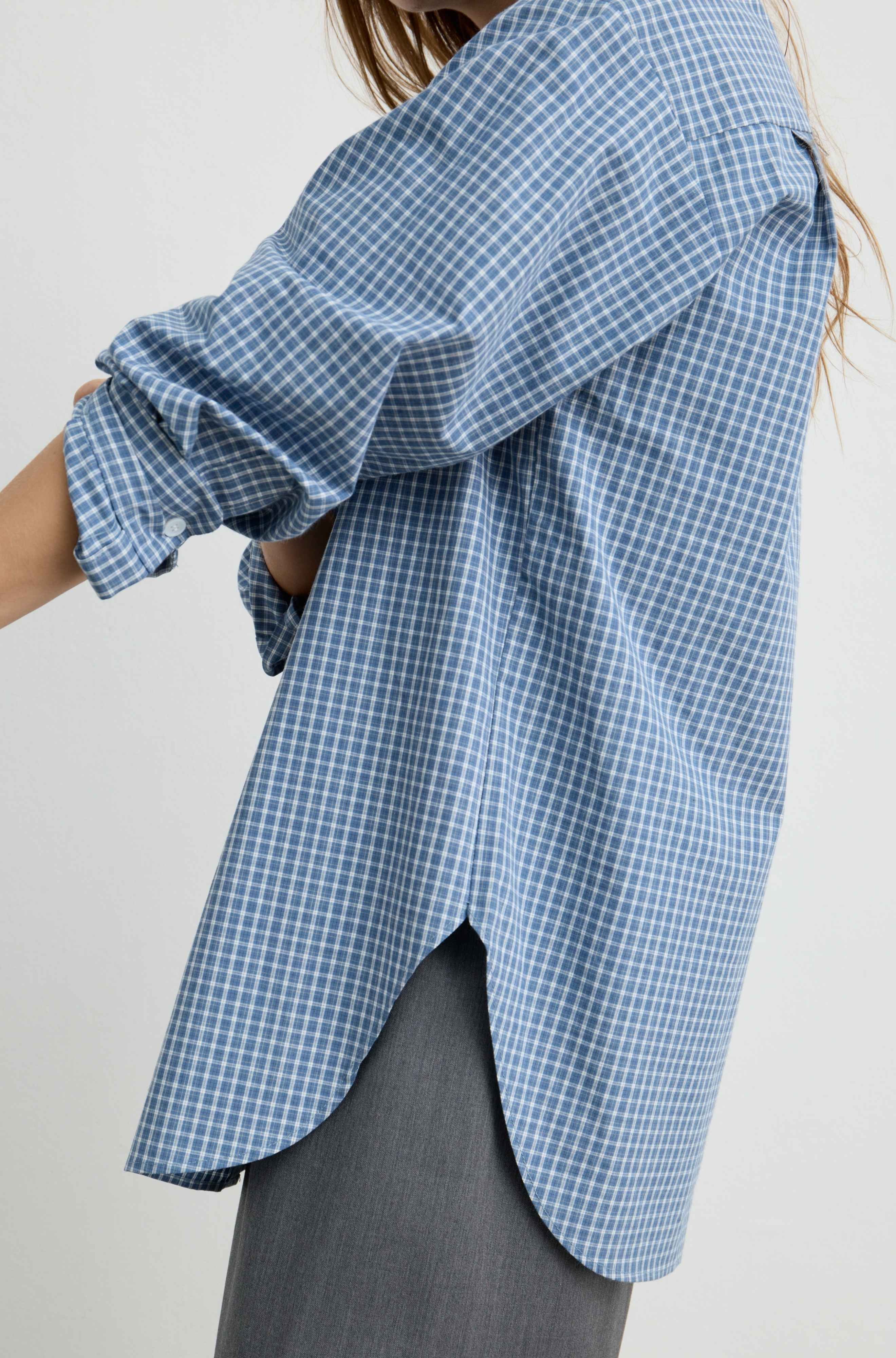 A close-up of a person wearing an oversized blue and white gingham check shirt with rolled sleeves and a curved hem, paired with gray pants against a plain light background.