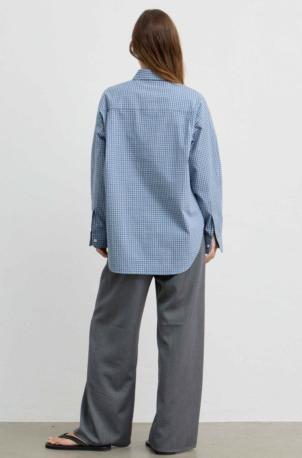A woman shown from the back wearing a light blue and white gingham long-sleeve button-up shirt and wide-leg gray trousers, standing against a plain light gray studio background.