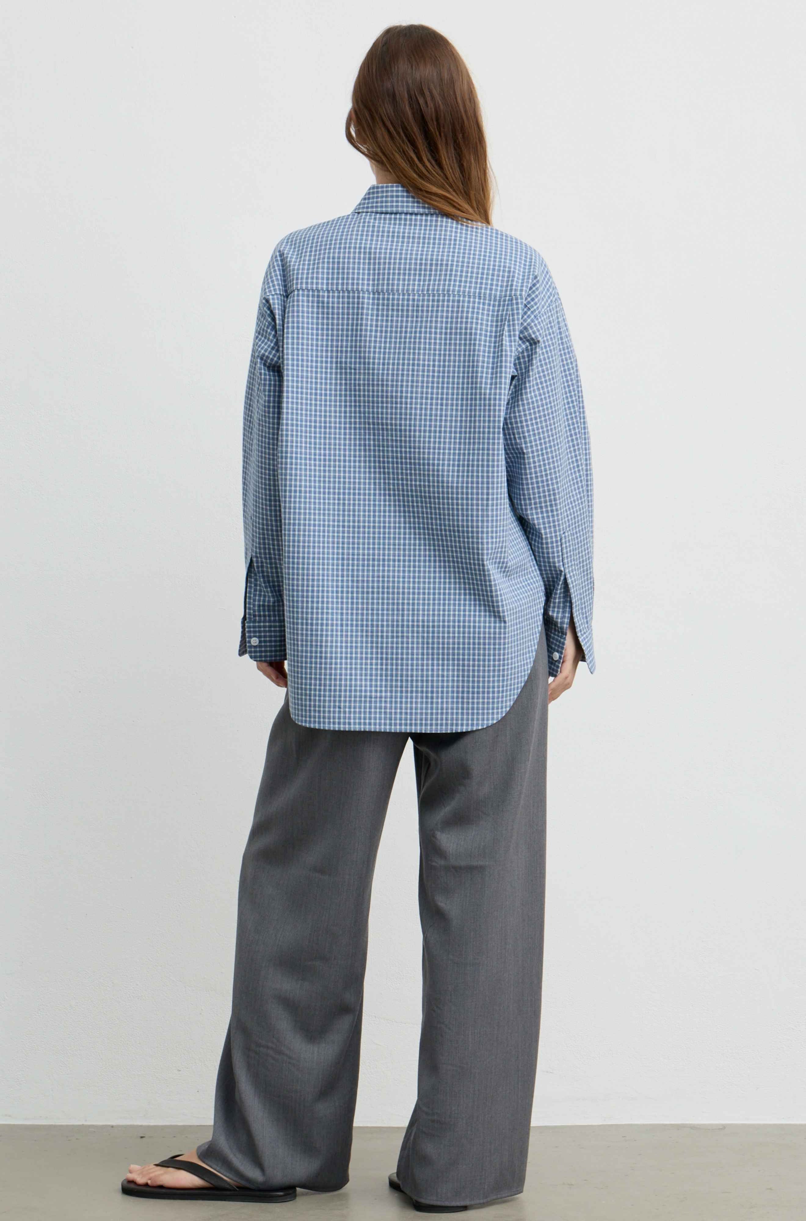 A woman shown from the back wearing a light blue and white gingham long-sleeve button-up shirt and wide-leg gray trousers, standing against a plain light gray studio background.