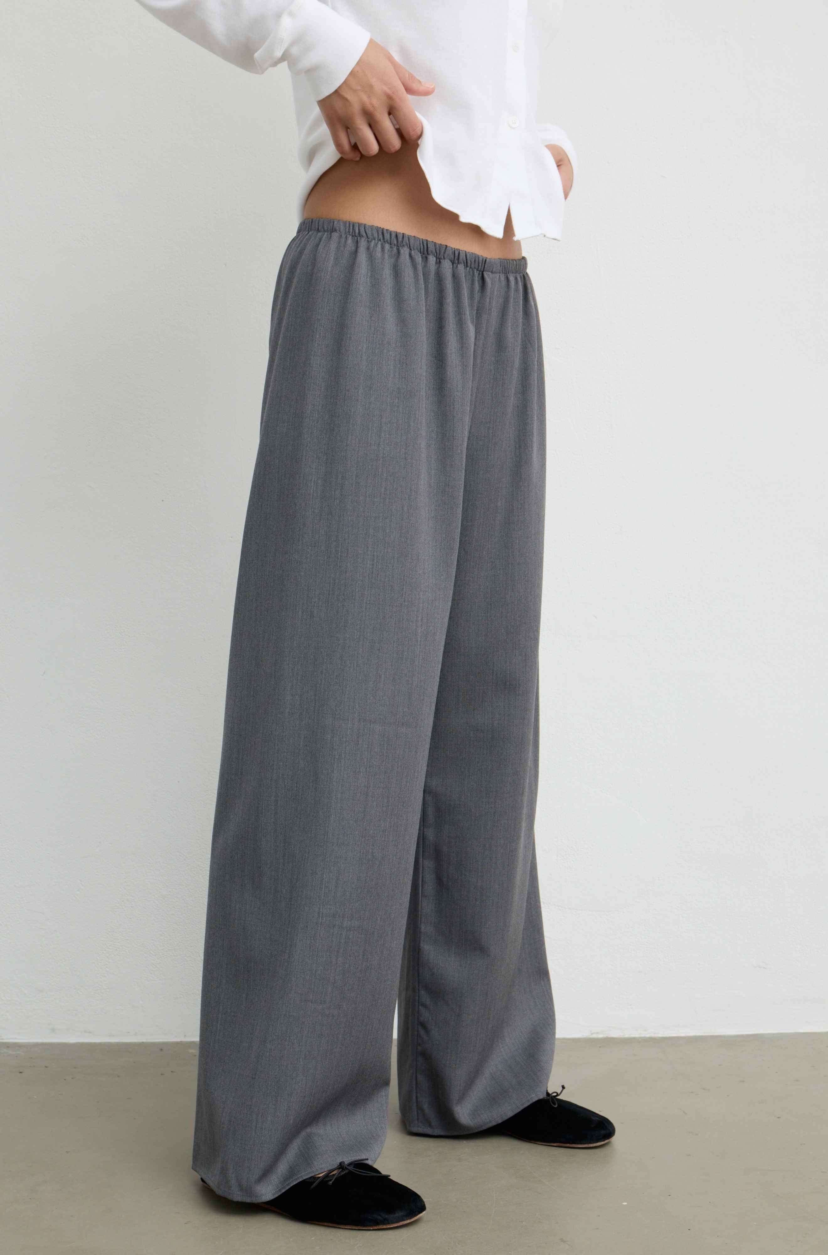 A person wearing loose, wide-leg gray pants with an elastic waistband, styled with a white shirt and black slip-on shoes against a plain light gray wall and concrete floor.