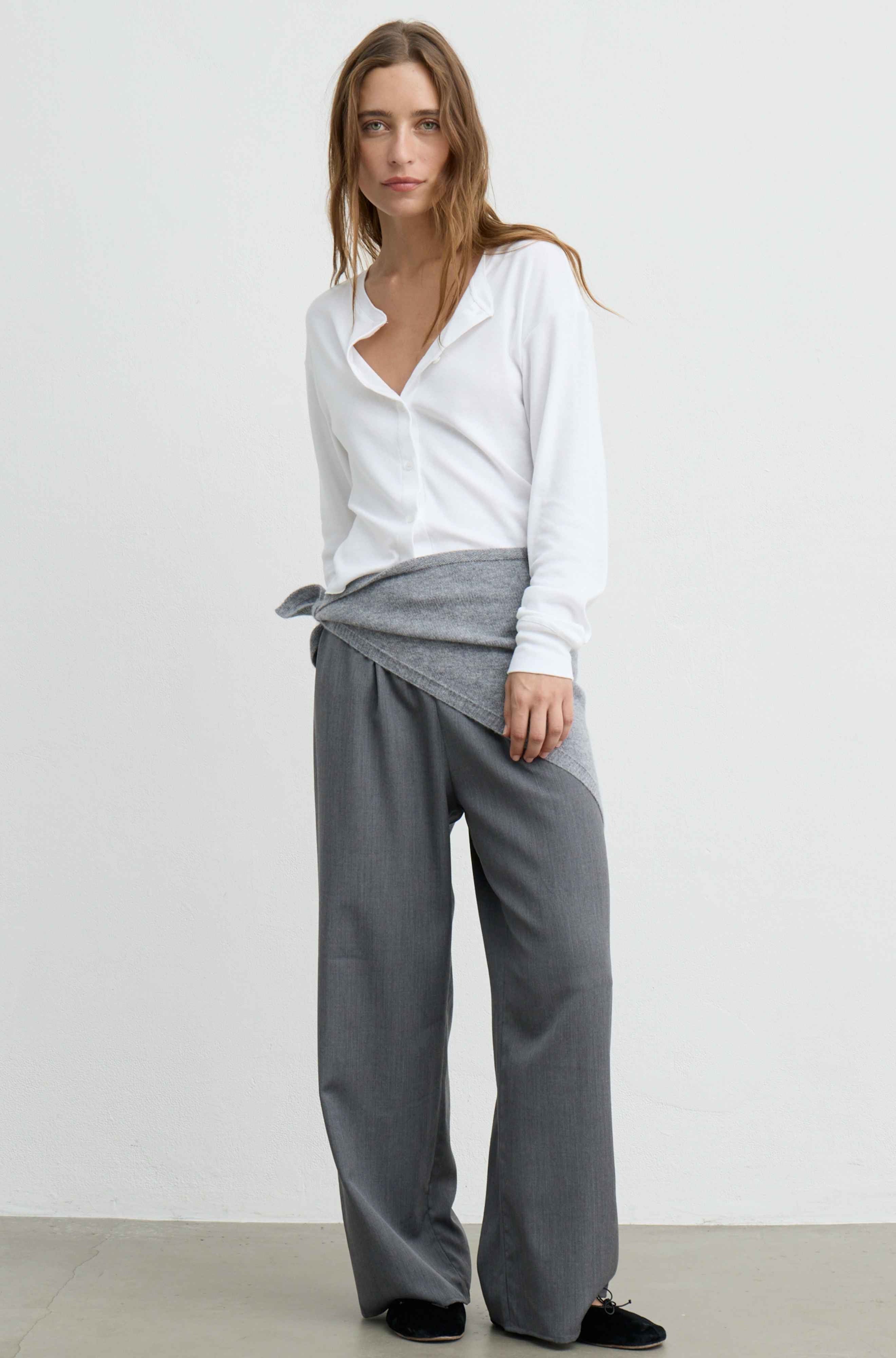 A woman modeling a white long-sleeve button-up shirt and wide-leg gray trousers, standing against a plain white studio wall.