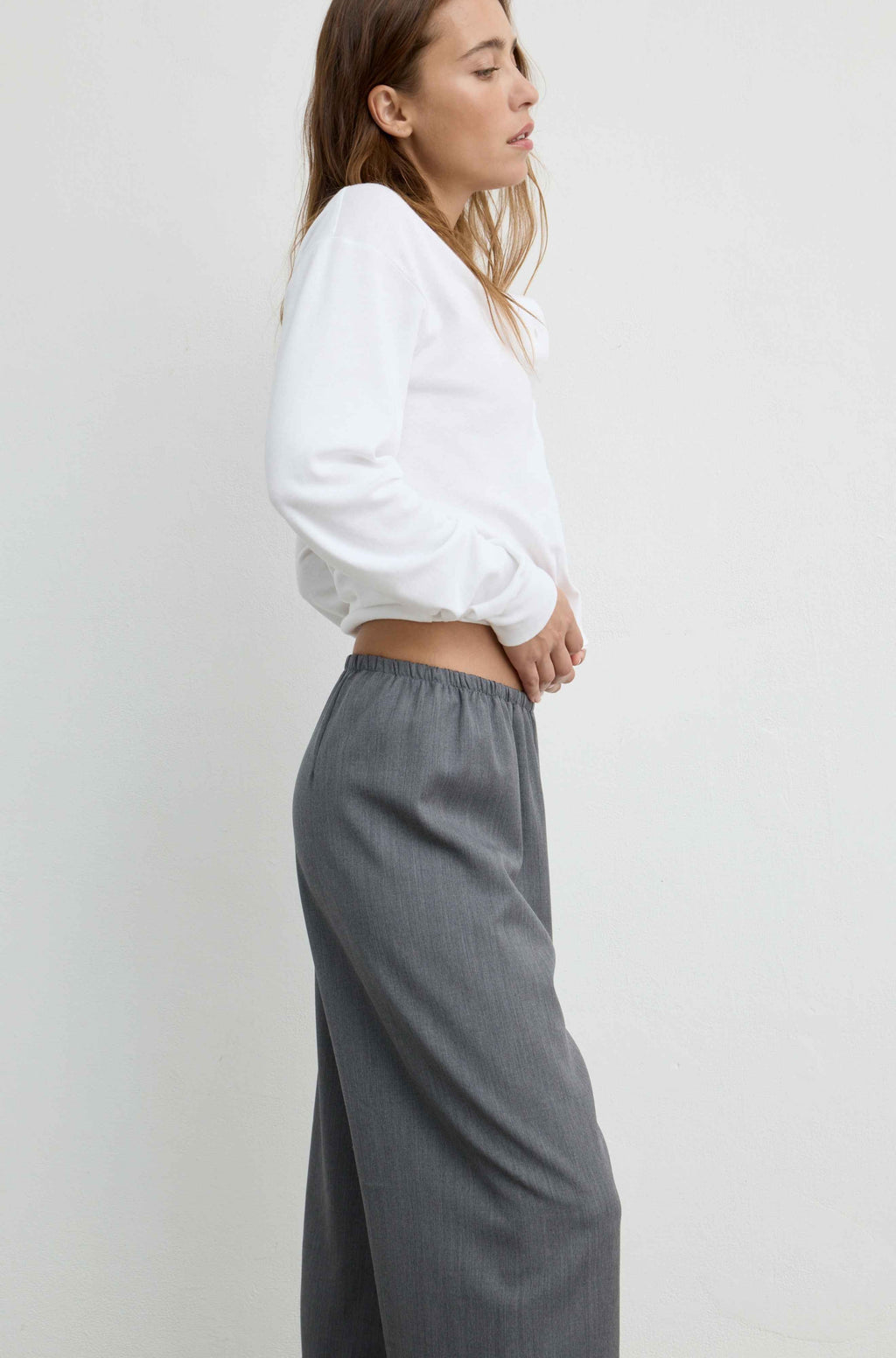 A woman shown in side profile wearing a white cropped long-sleeve top and loose gray wide-leg trousers against a plain light gray studio background.