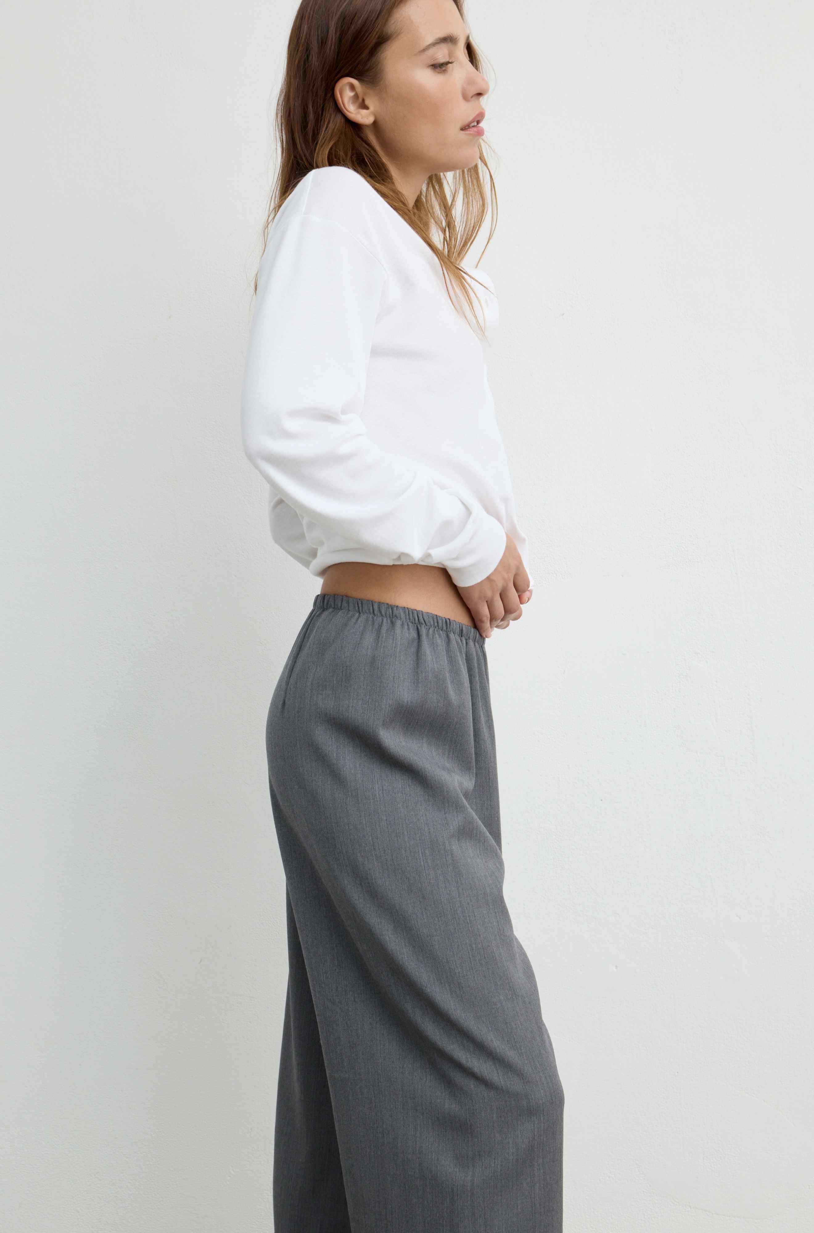 A woman shown in side profile wearing a white cropped long-sleeve top and loose gray wide-leg trousers against a plain light gray studio background.