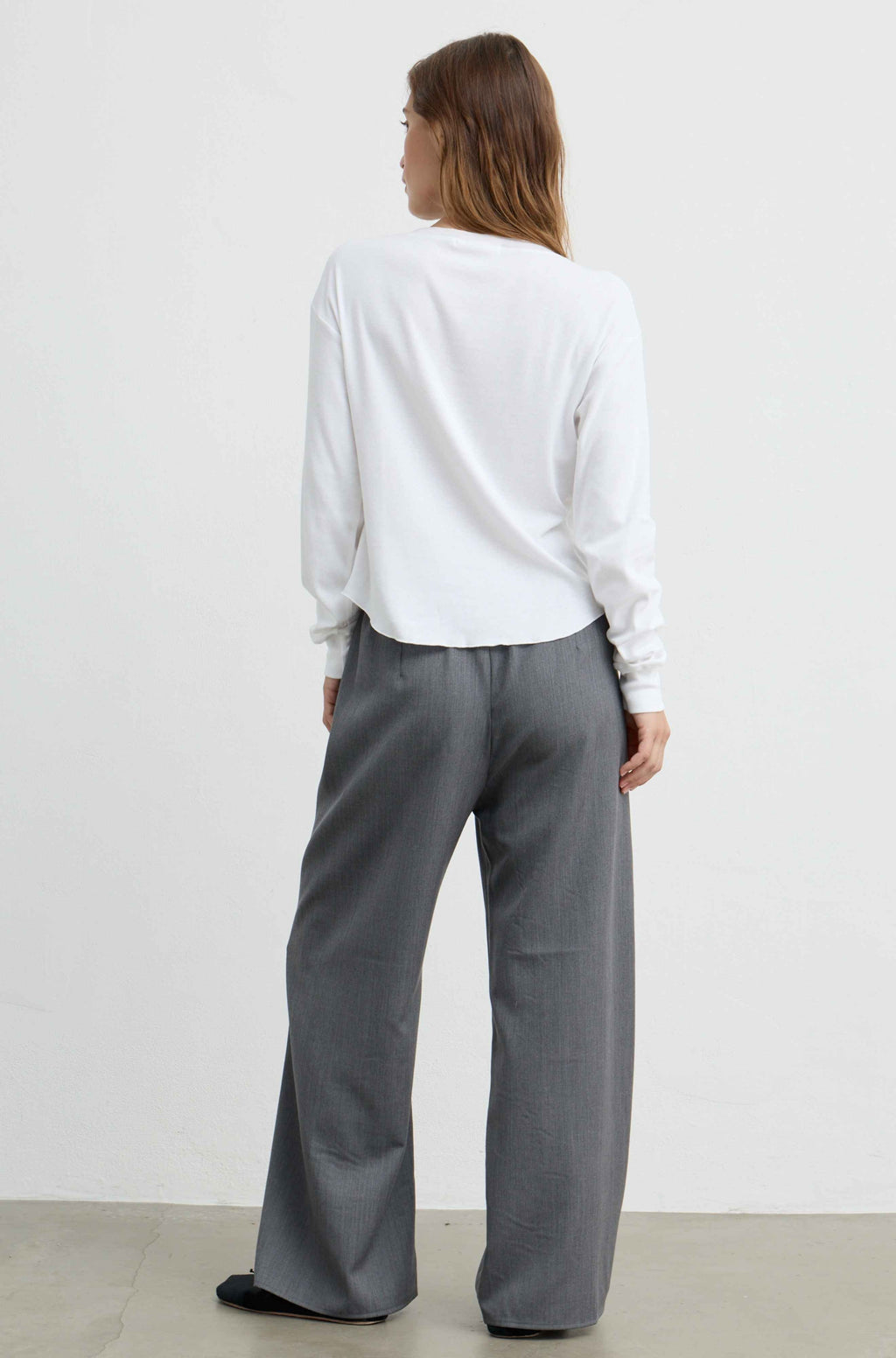 A woman shown from the back wearing a white long-sleeve top and gray wide-leg trousers, standing against a plain light gray wall.