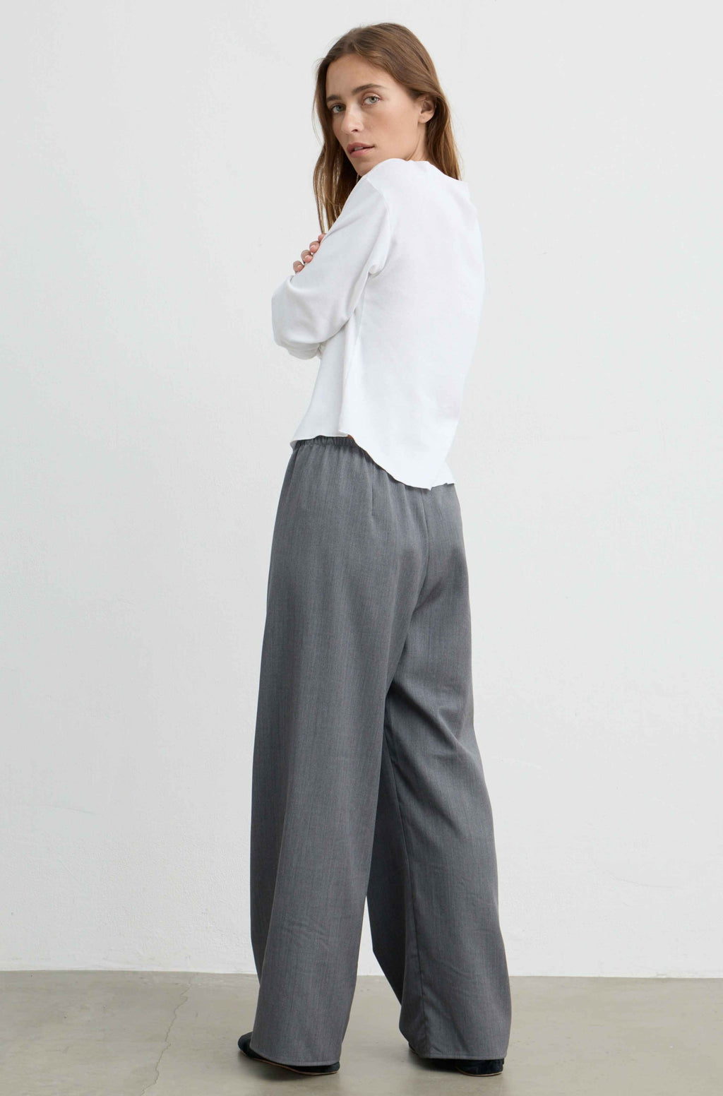 A woman wearing a white long-sleeve blouse and gray wide-leg trousers, photographed from the back in a minimalist studio setting.