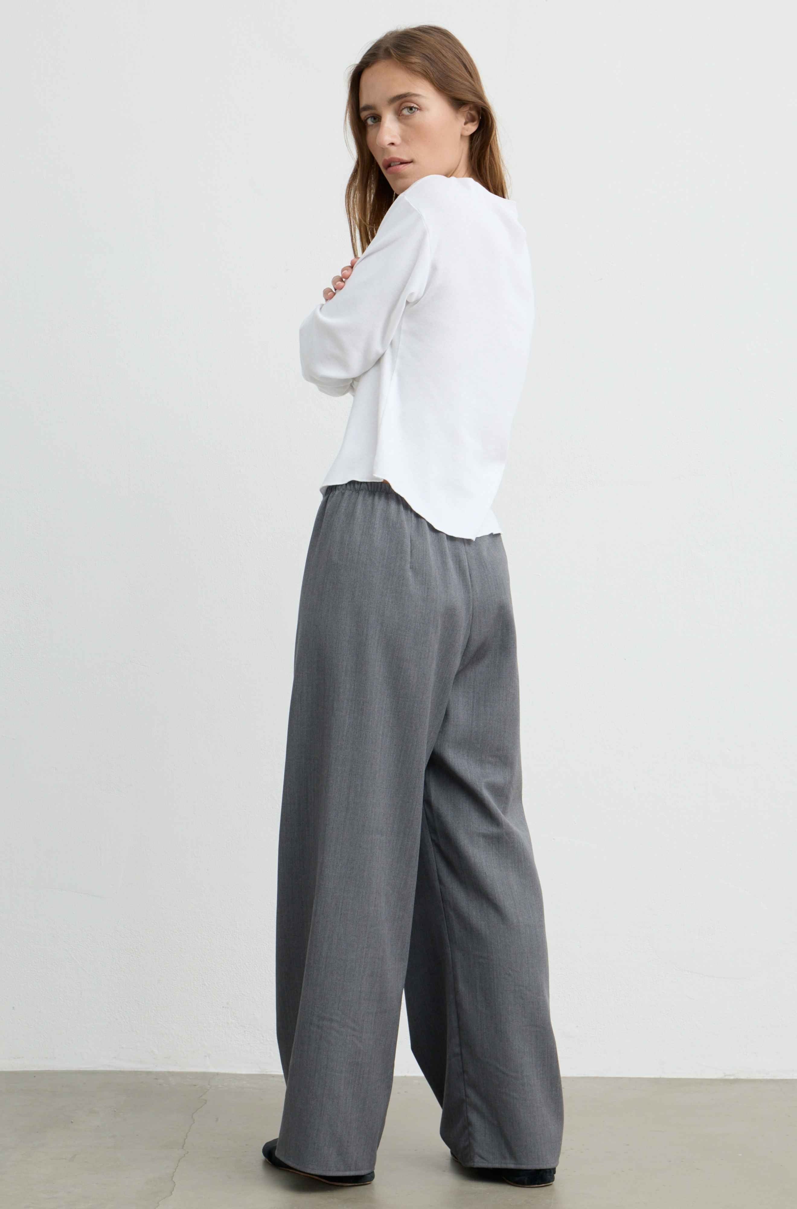 A woman wearing a white long-sleeve blouse and gray wide-leg trousers, photographed from the back in a minimalist studio setting.