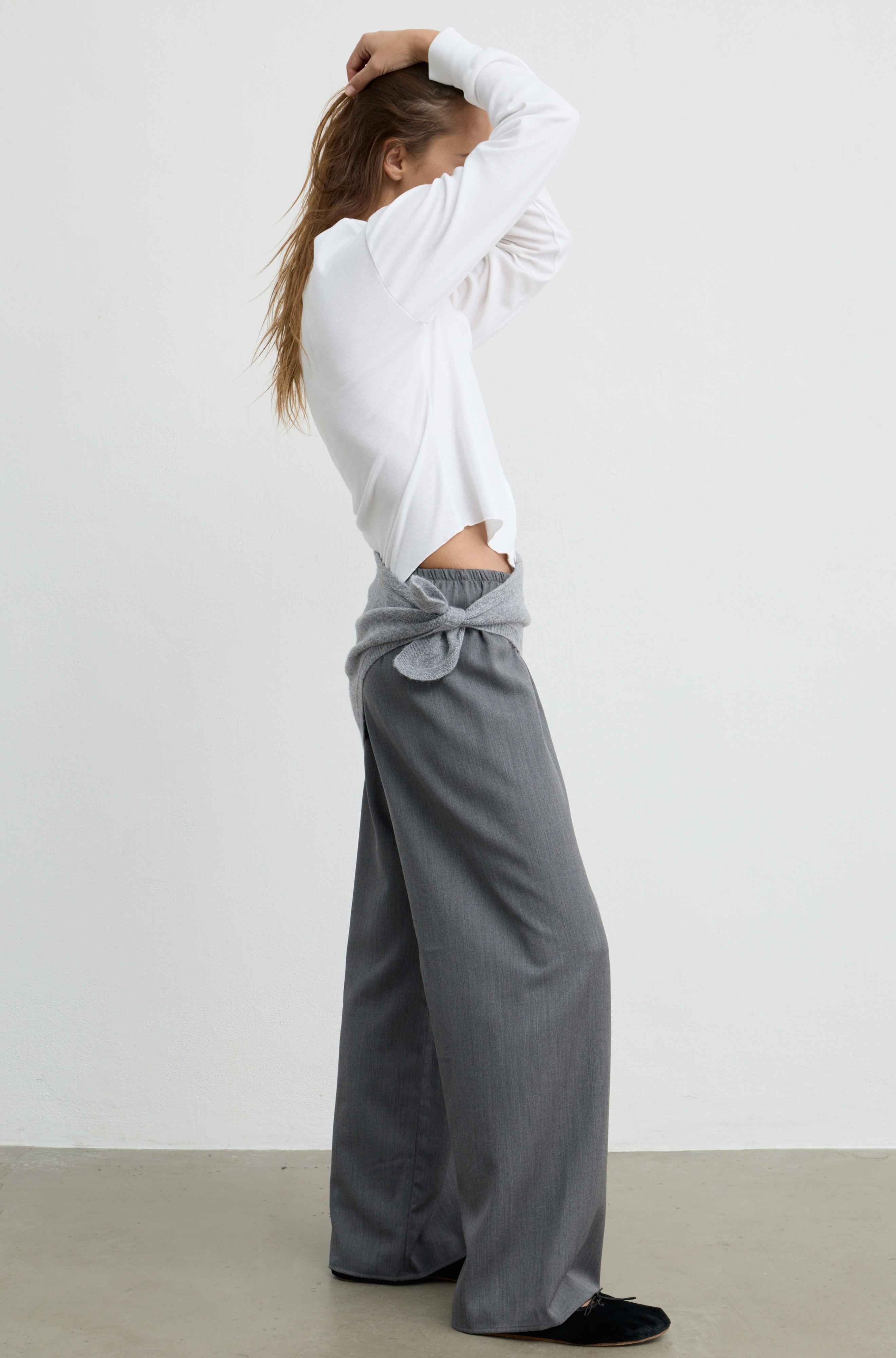 A woman stands in profile wearing a white long-sleeve top and gray wide-leg pants tied at the waist, photographed against a plain light gray wall and concrete floor.