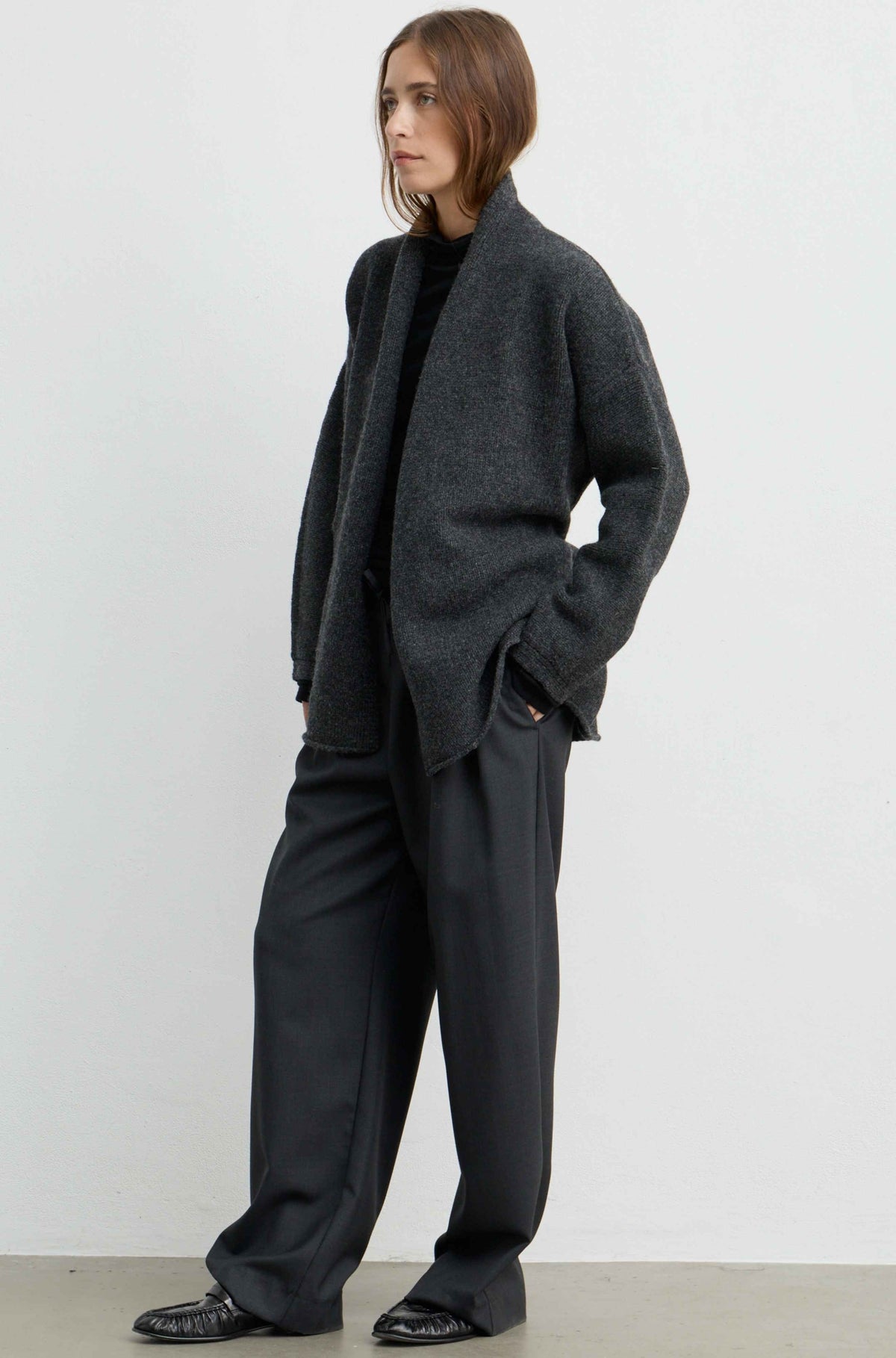 A model wearing a dark gray open-front knit cardigan, black wide-leg trousers, and black shoes, standing against a plain white studio wall.