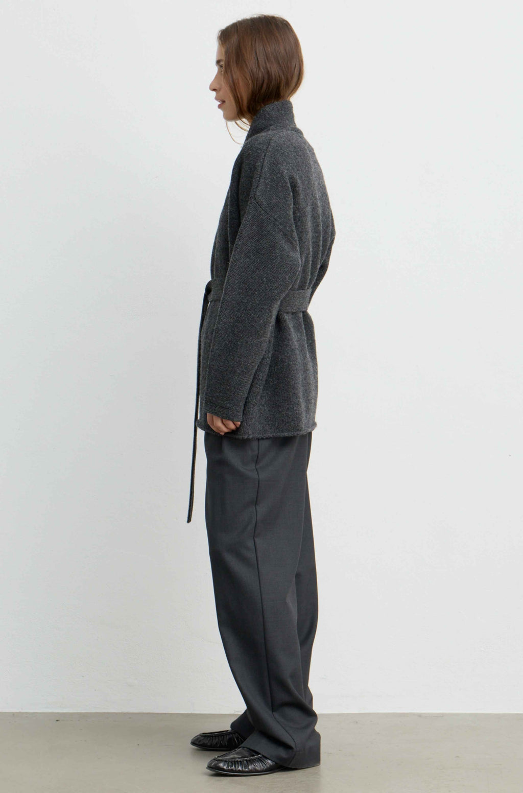 A model shown from the side profile wearing a dark gray textured wrap jacket and matching dark gray wide-leg trousers against a plain white studio background.