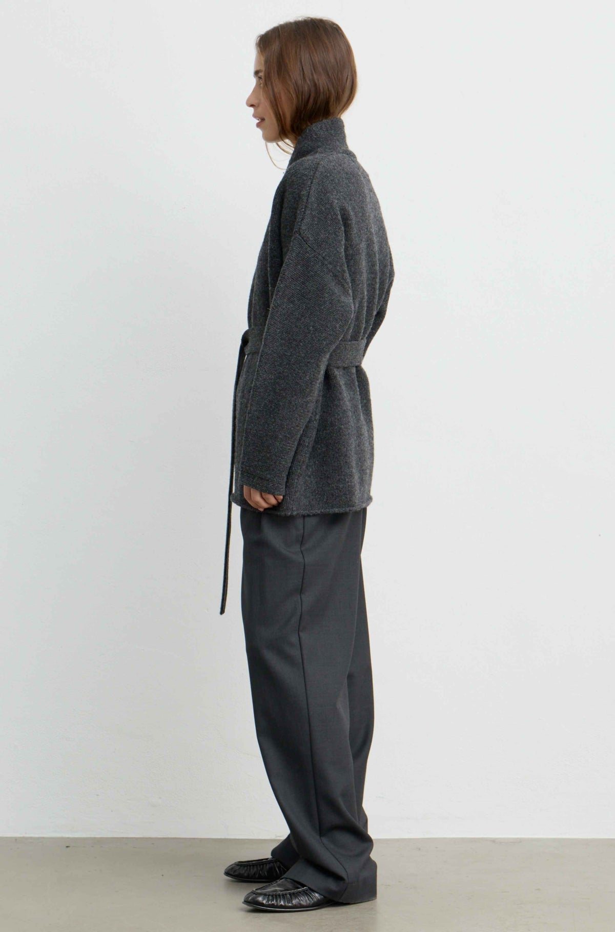 A model shown from the side profile wearing a dark gray textured wrap jacket and matching dark gray wide-leg trousers against a plain white studio background.