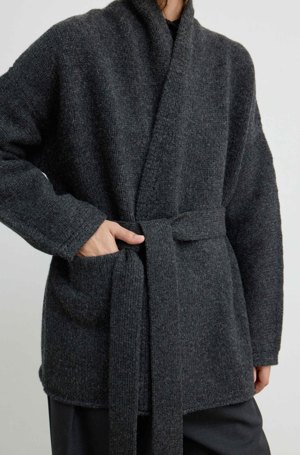 A close-up of a person wearing a dark charcoal gray textured knit wrap coat with a matching tie belt and front patch pockets, paired with dark trousers against a light gray studio background.