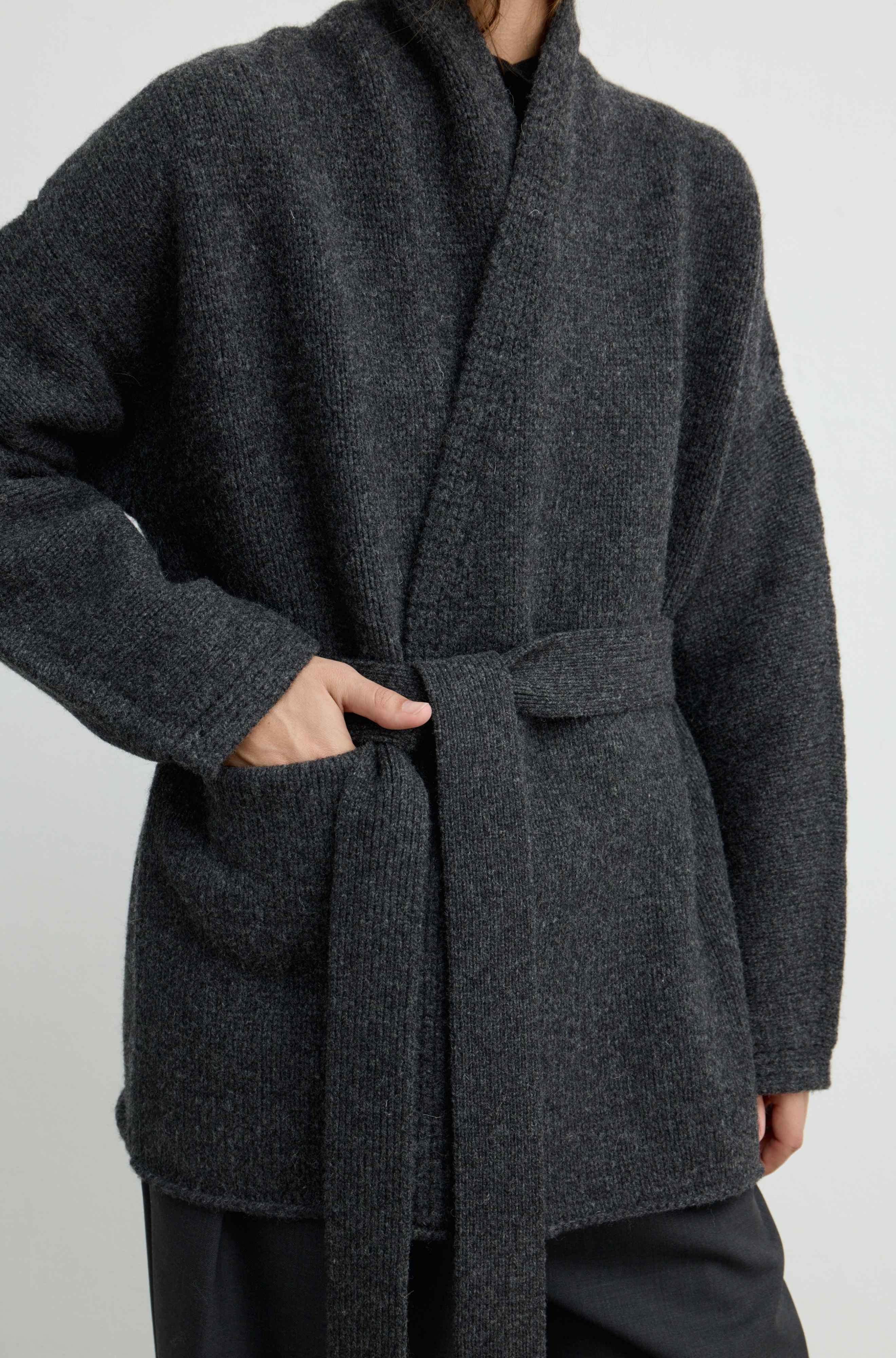 A close-up of a person wearing a dark charcoal gray textured knit wrap coat with a matching tie belt and front patch pockets, paired with dark trousers against a light gray studio background.