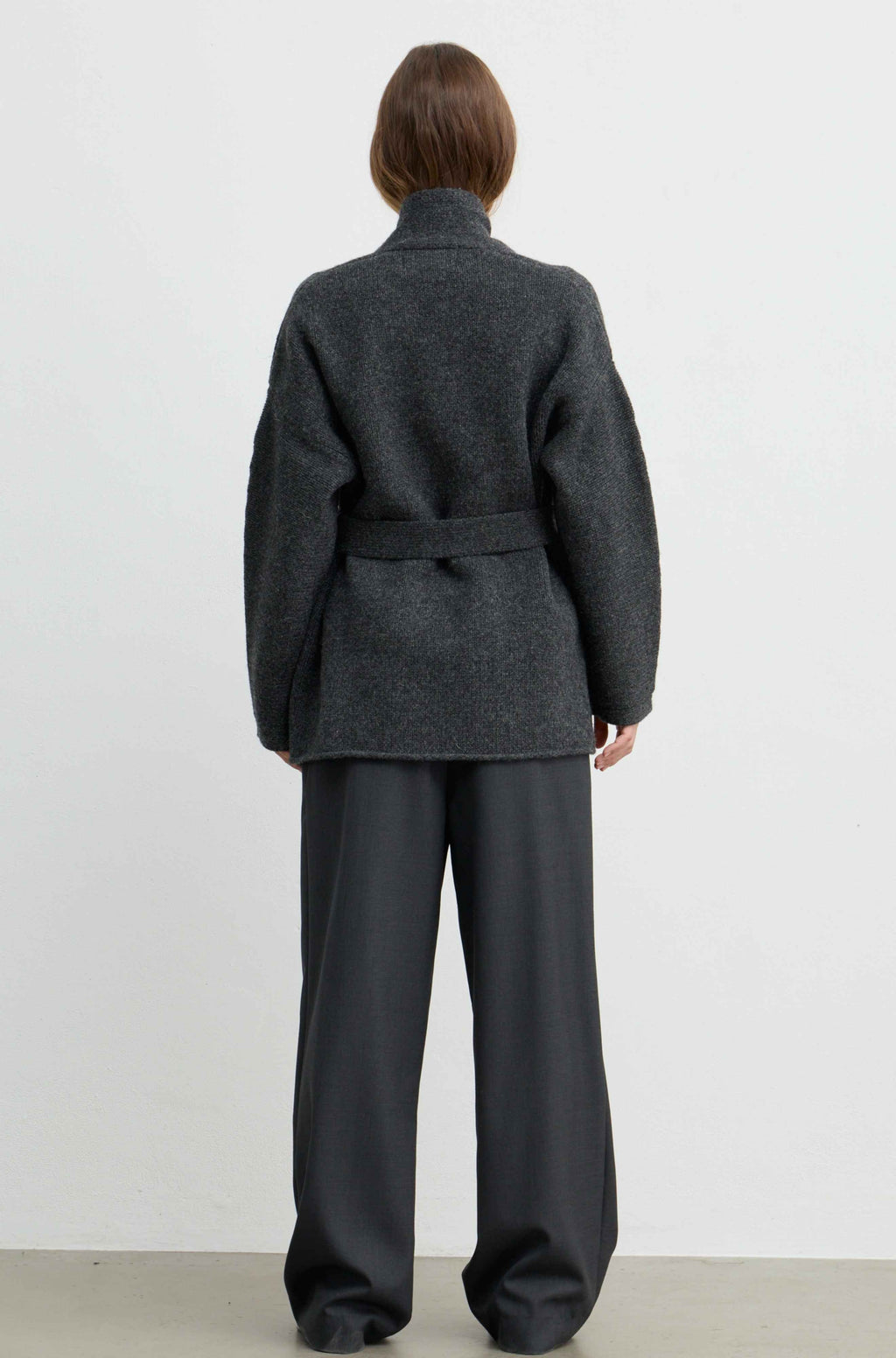 A model viewed from the back wearing a dark charcoal gray belted coat and matching wide-leg trousers against a plain white studio background.