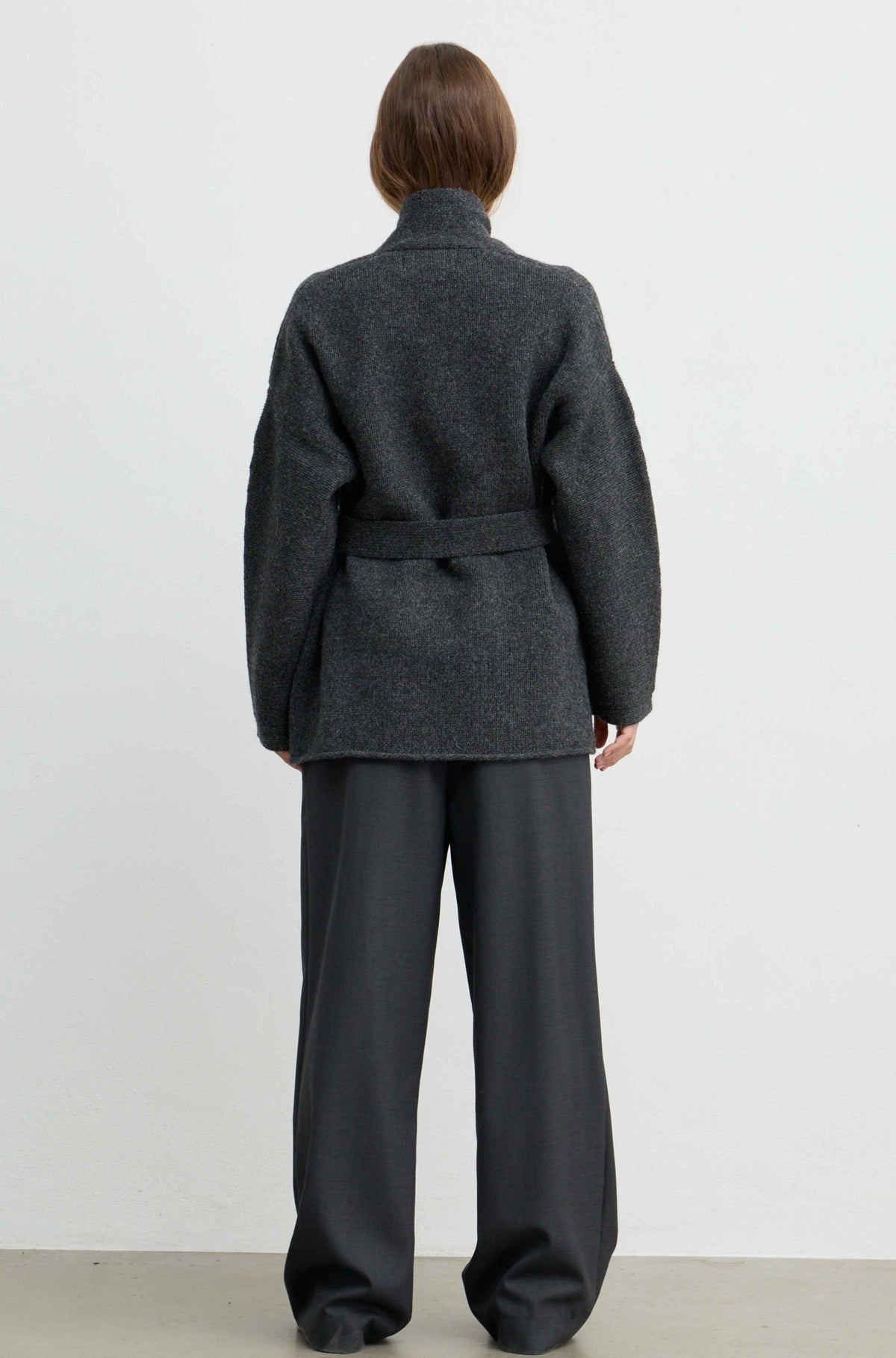 A model viewed from the back wearing a dark charcoal gray belted coat and matching wide-leg trousers against a plain white studio background.