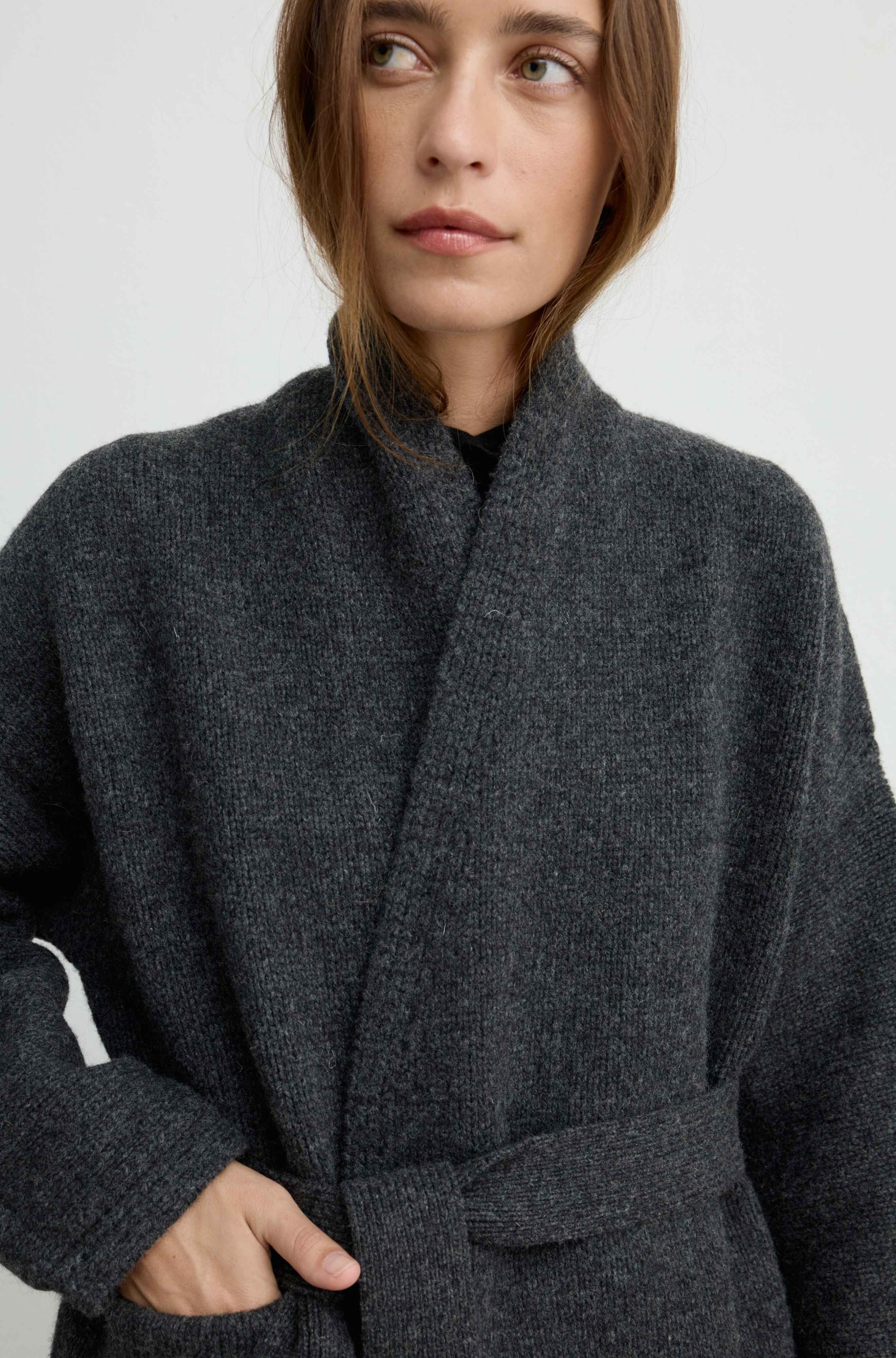 A close-up of a woman wearing a dark charcoal gray textured wrap coat with a high collar and a tie belt, set against a plain light gray background.