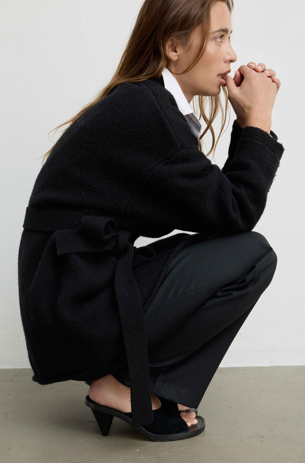 A woman crouches in a studio wearing a black belted cardigan, black wide-leg trousers, and black heeled sandals over a white collared shirt.