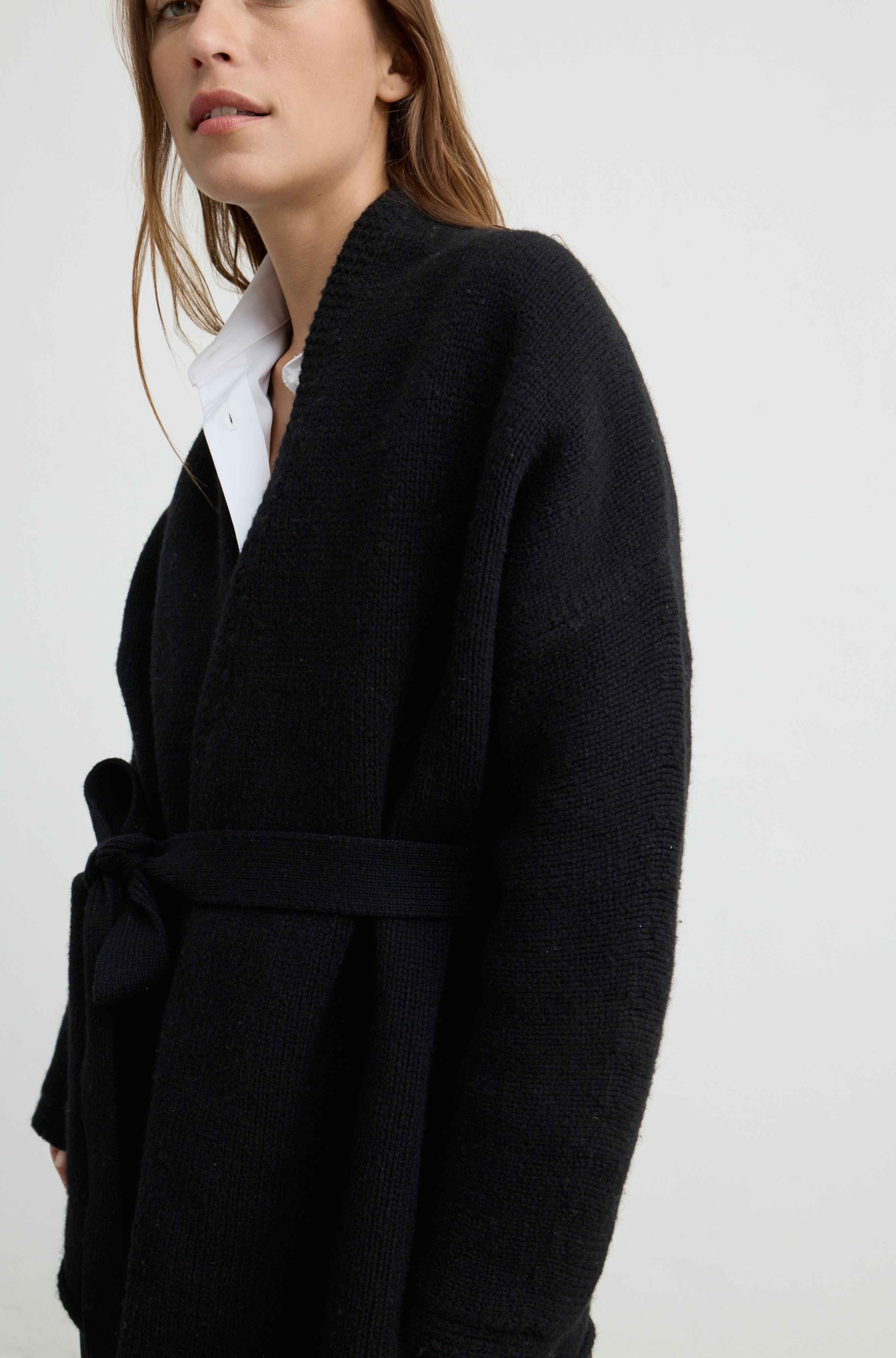 A close-up of a model wearing a black textured knit wrap coat with a matching tie belt over a white collared shirt, set against a plain light gray background.