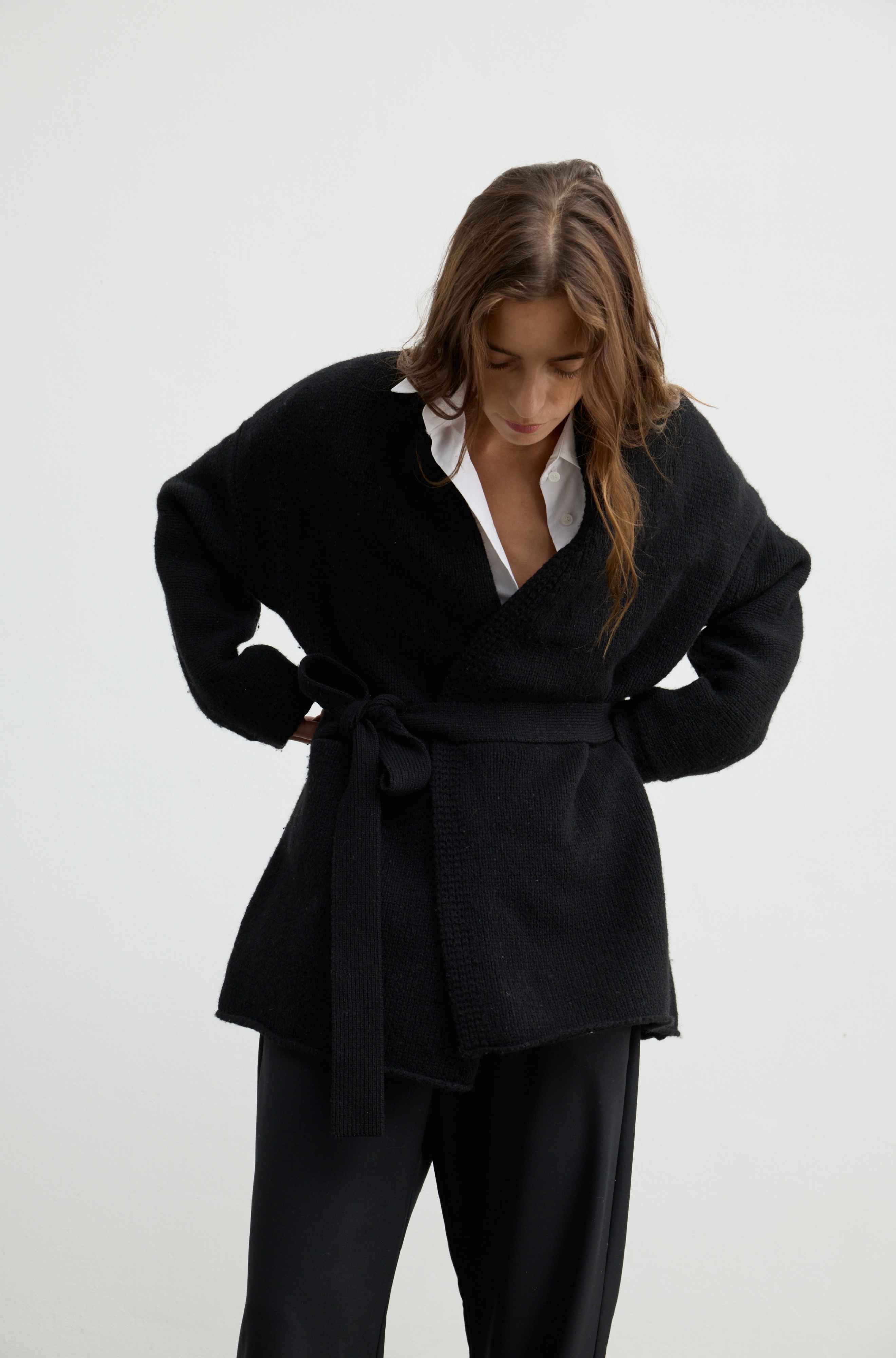 A woman wearing a black textured knit wrap-style cardigan with a tie belt over a white collared shirt and black trousers, photographed against a plain light gray studio background.