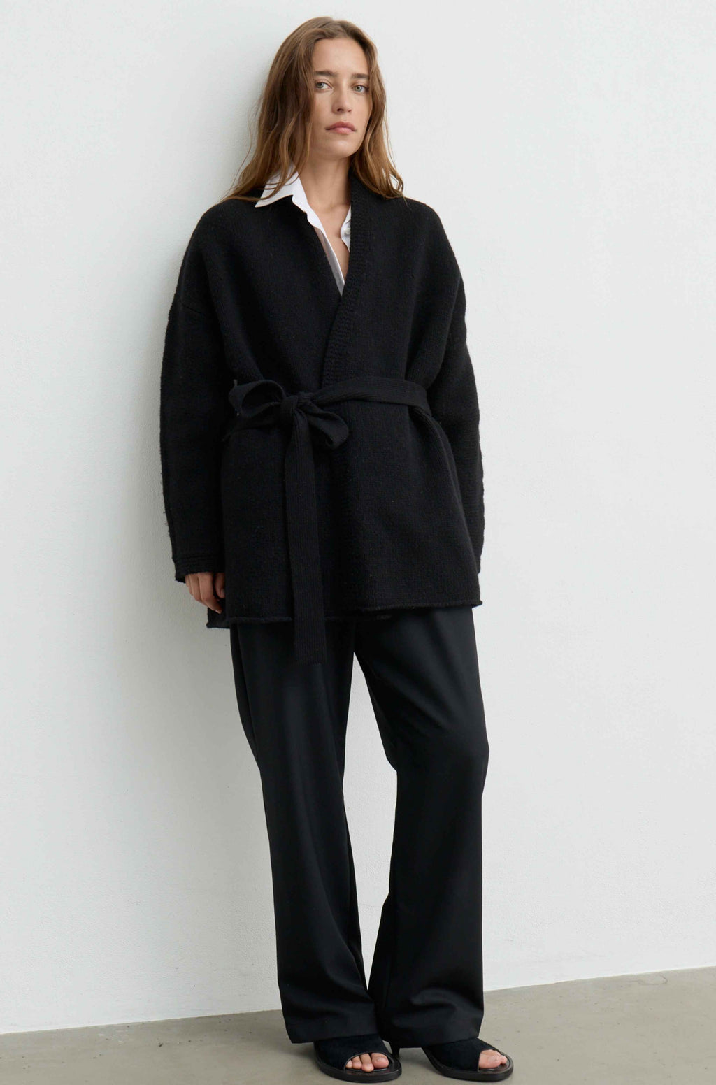A woman stands against a plain light gray wall wearing an oversized black belted cardigan or wrap jacket over a white collared shirt, with loose black wide-leg pants and black slide sandals.