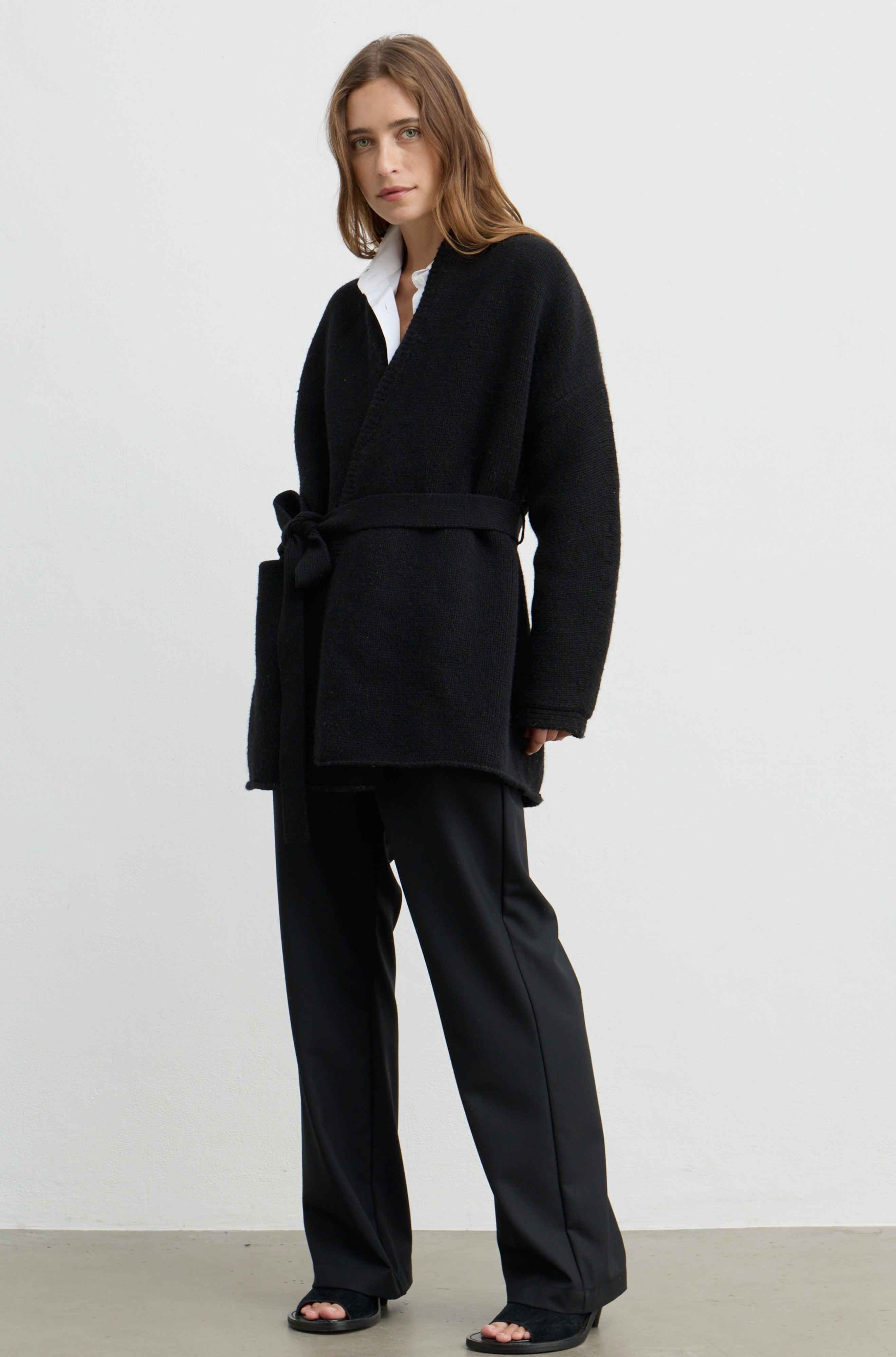 A woman modeling a black belted wrap coat over a white collared shirt and black wide-leg trousers, standing against a plain light gray studio background.