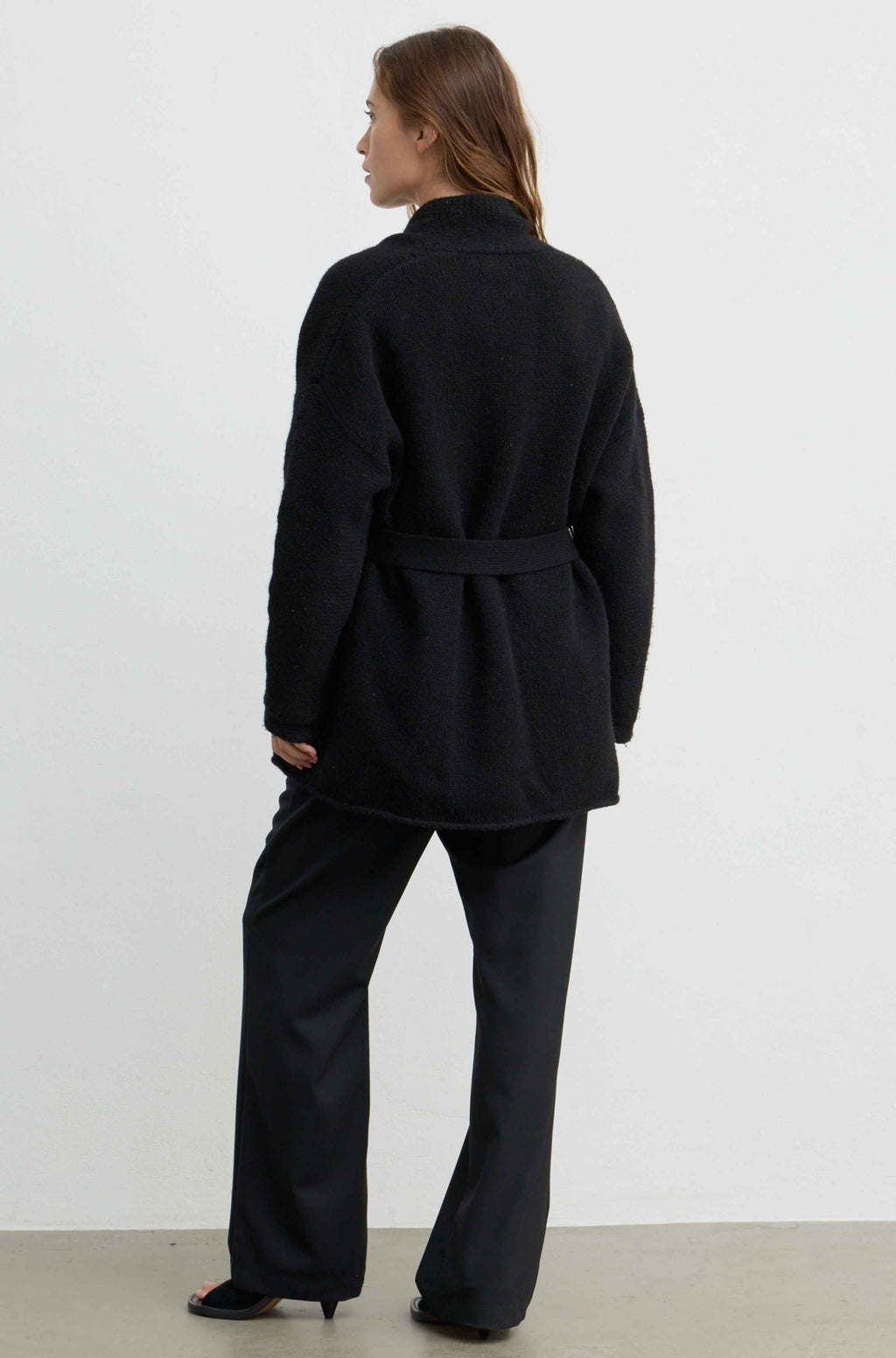 A woman shown from the back wearing a black belted coat and black wide-leg trousers against a plain light gray studio background.