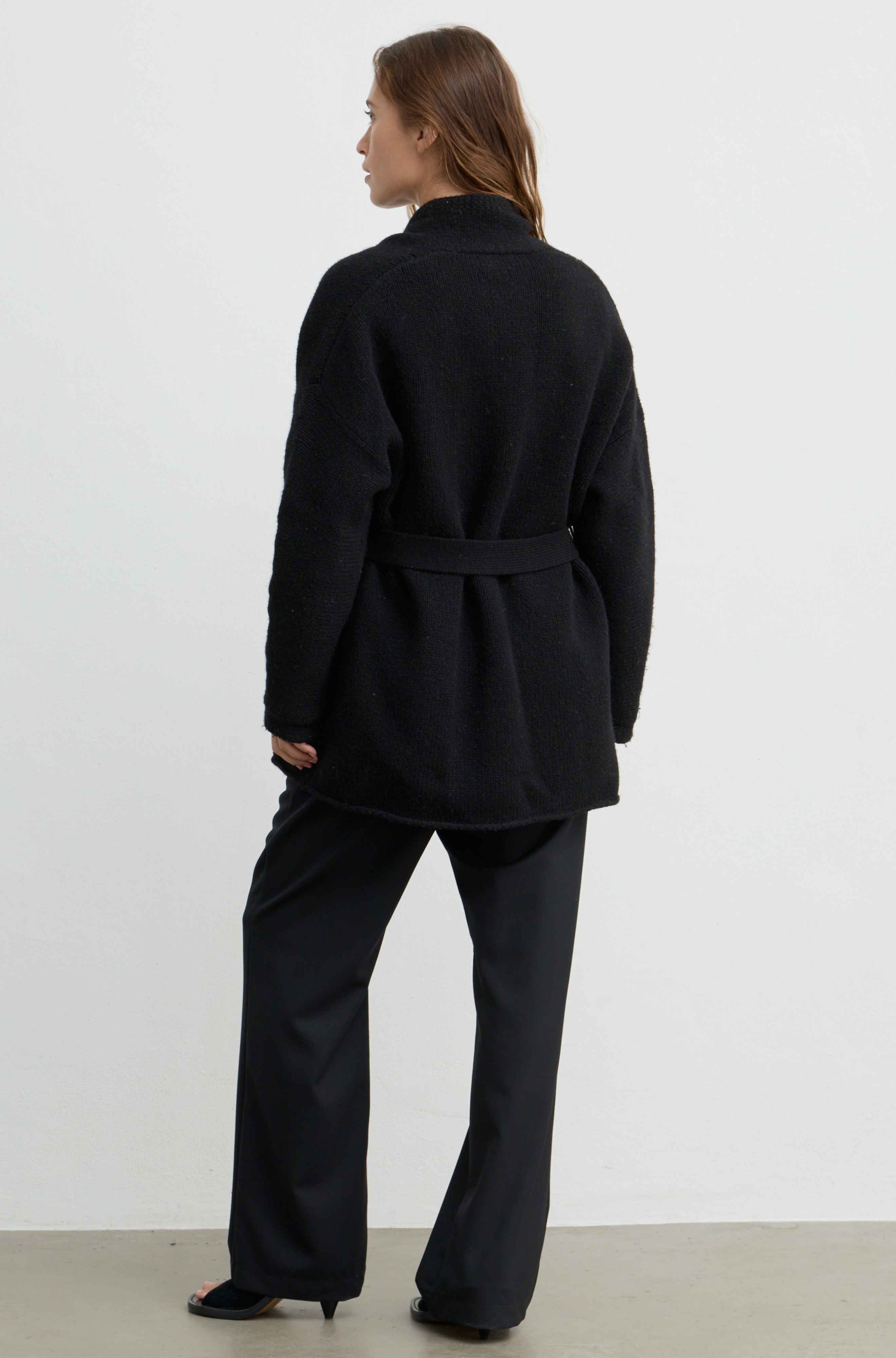 A woman shown from the back wearing a black belted coat and black wide-leg trousers against a plain light gray studio background.
