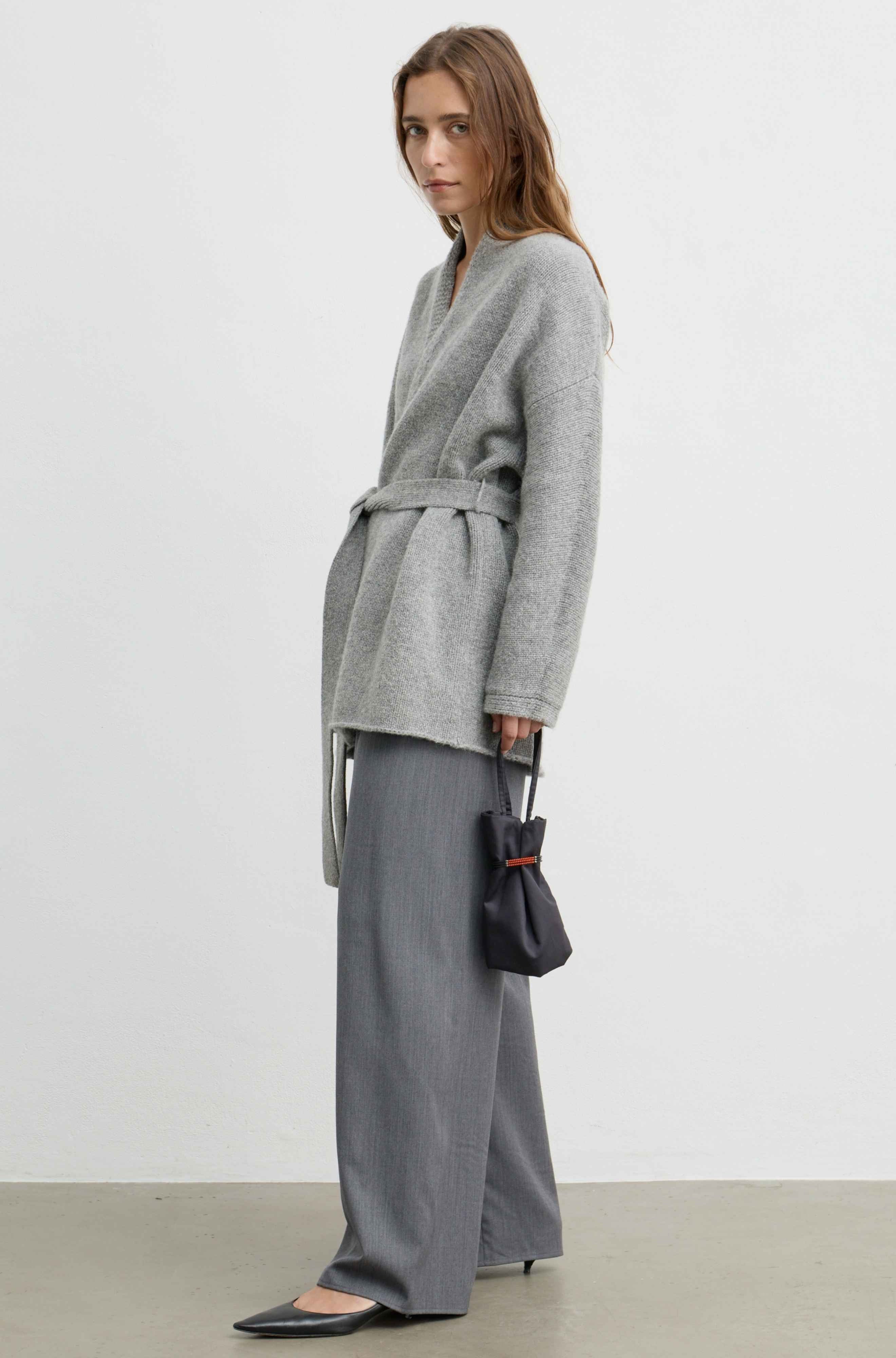 A woman stands in profile wearing a light gray belted cardigan, wide-leg gray trousers, and black pointed flats, while holding a small black handbag against a plain light gray studio background.