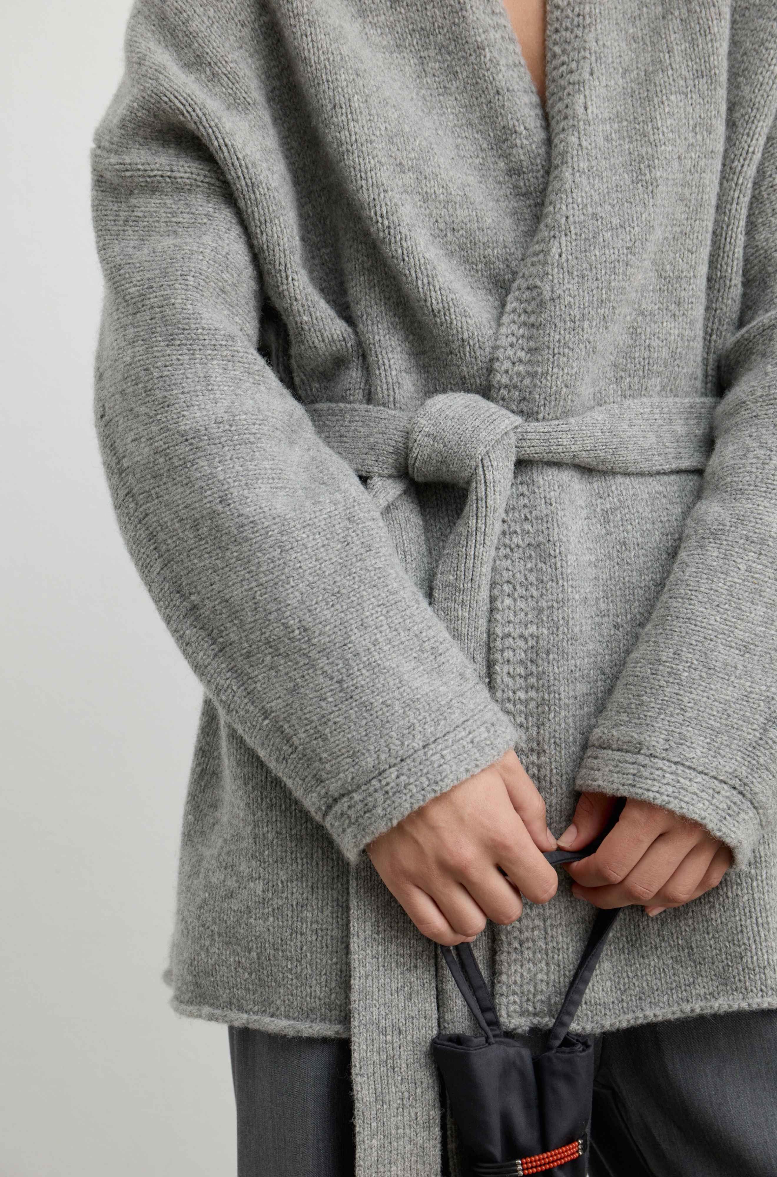 A close-up of a person wearing a chunky knit gray belted cardigan, holding a small black drawstring pouch.