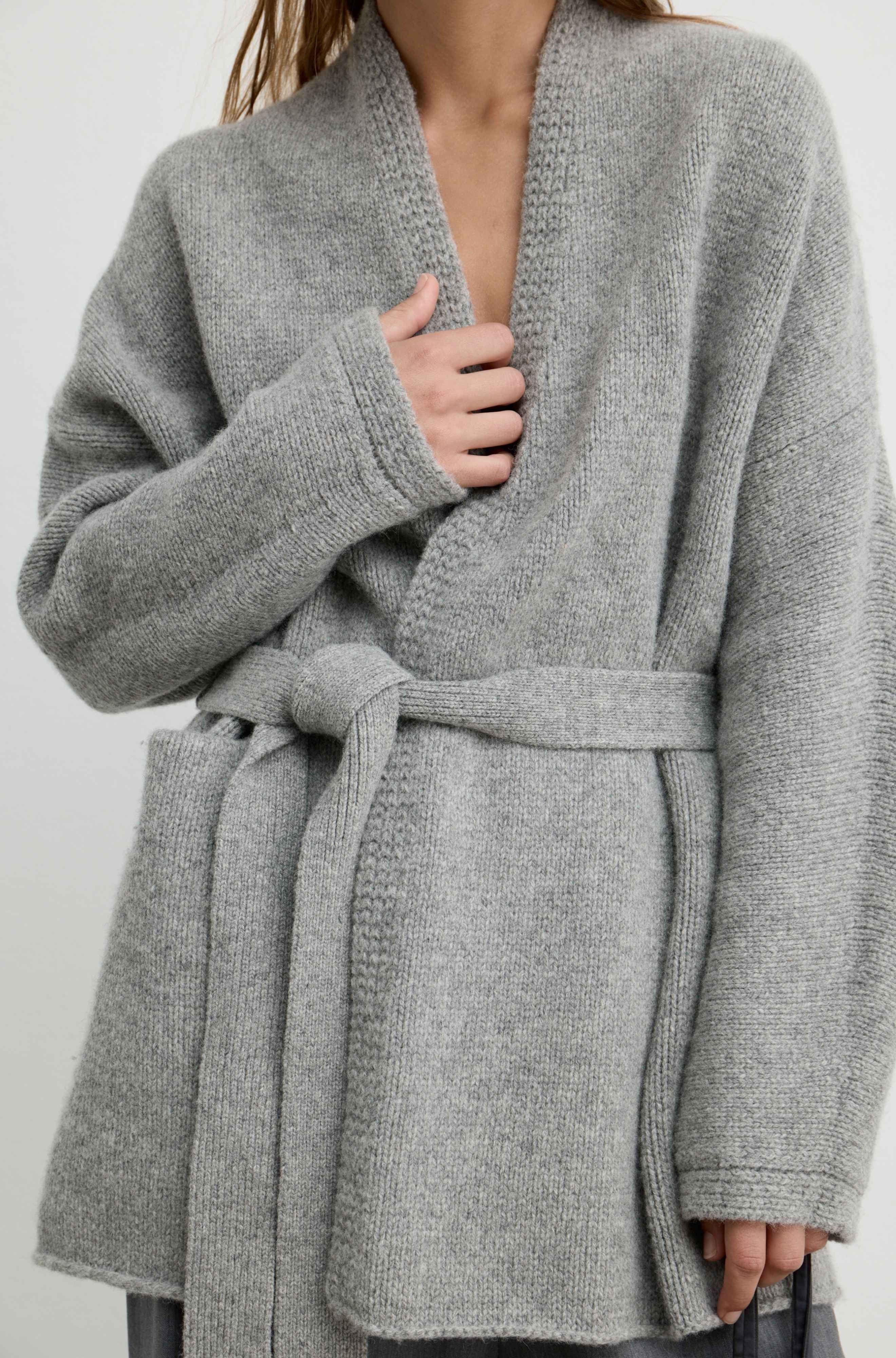 A close-up of a person wearing a light gray knit wrap cardigan with a matching tie belt, shown against a plain light background.