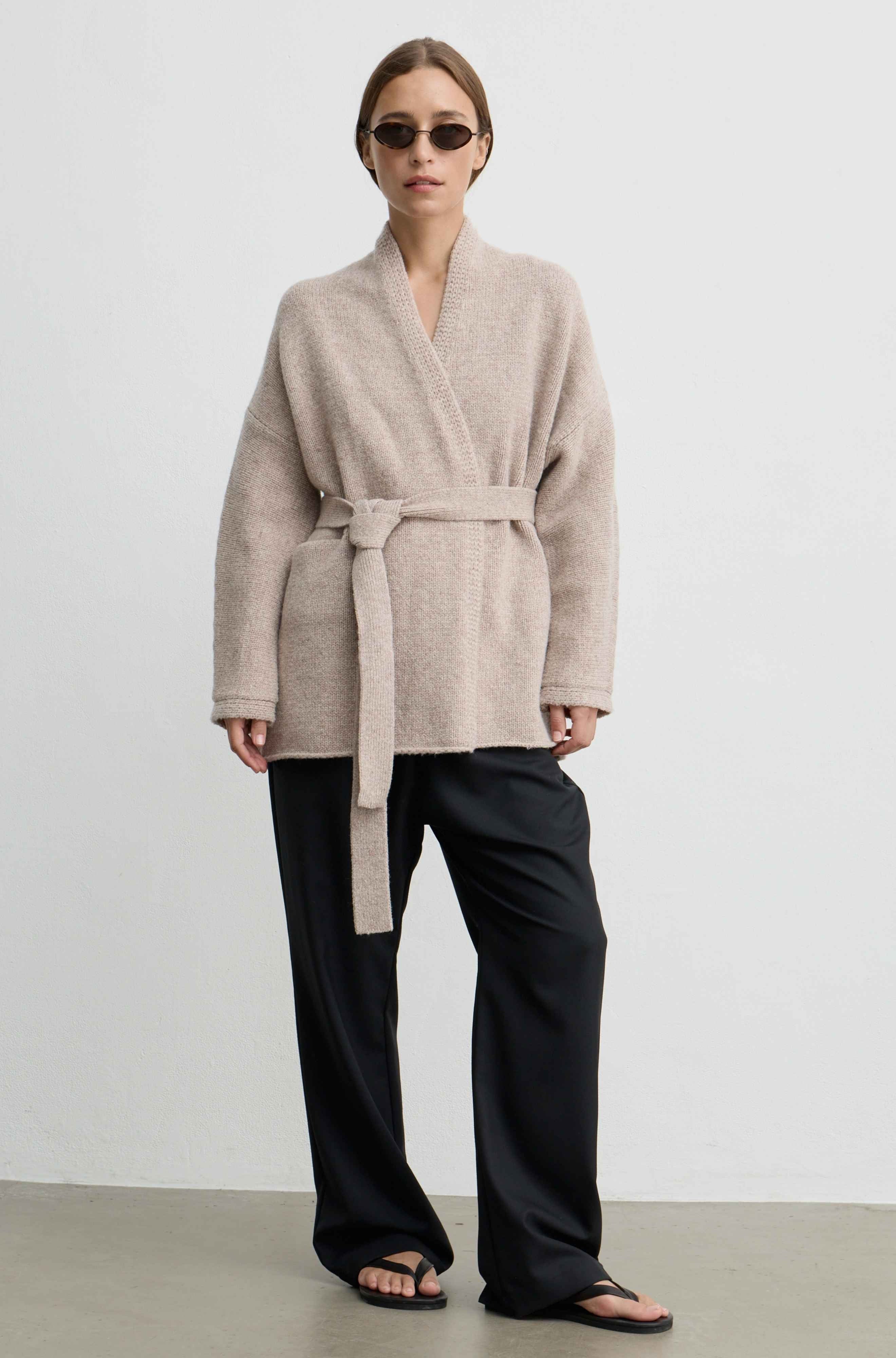 A model wearing a light beige knit wrap cardigan with a matching tie belt, paired with black wide-leg trousers and black sandals, standing against a plain light gray wall.