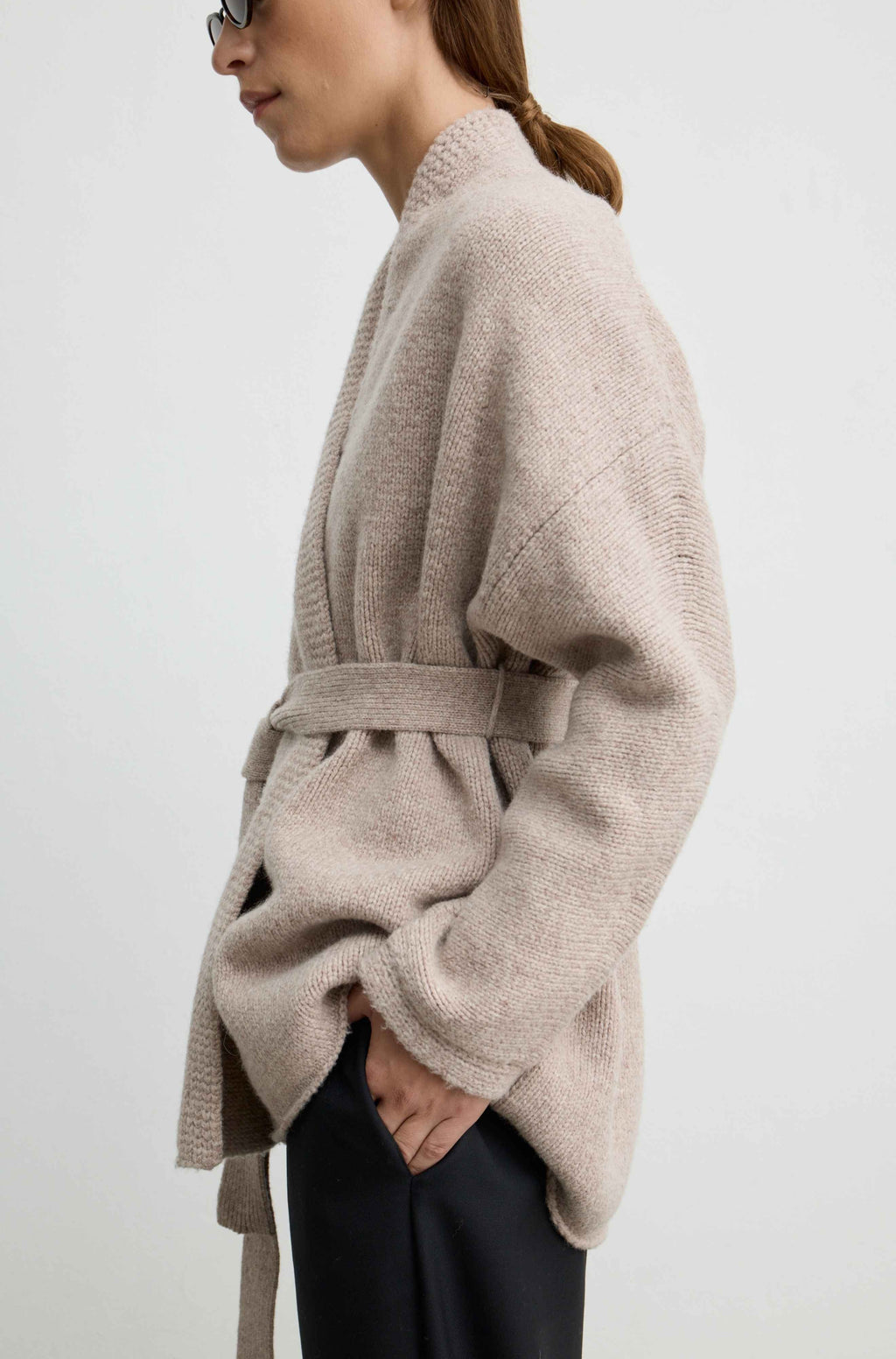 A woman shown in side profile wearing a light taupe knit belted cardigan and dark trousers against a plain light gray studio background.