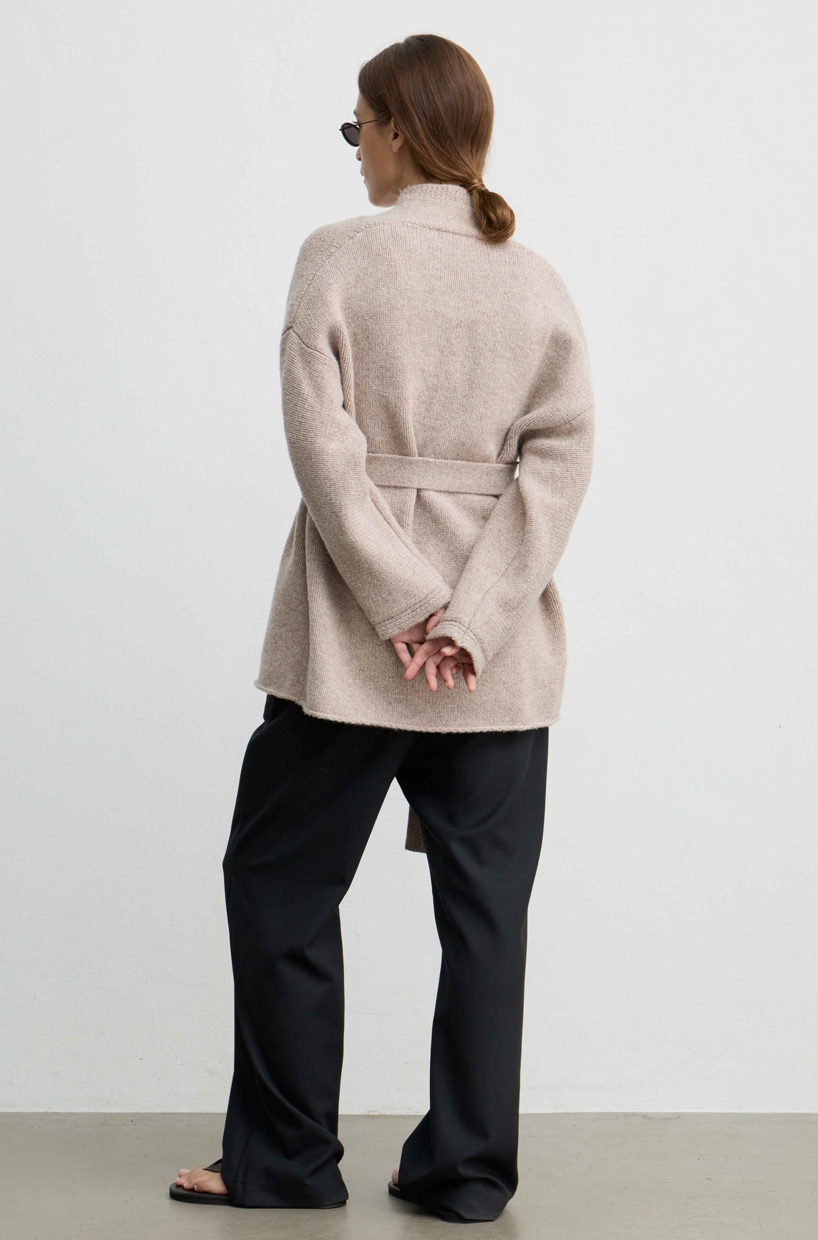 A woman shown from the back wearing a light beige belted knit cardigan and black wide-leg trousers, standing against a plain light gray wall.