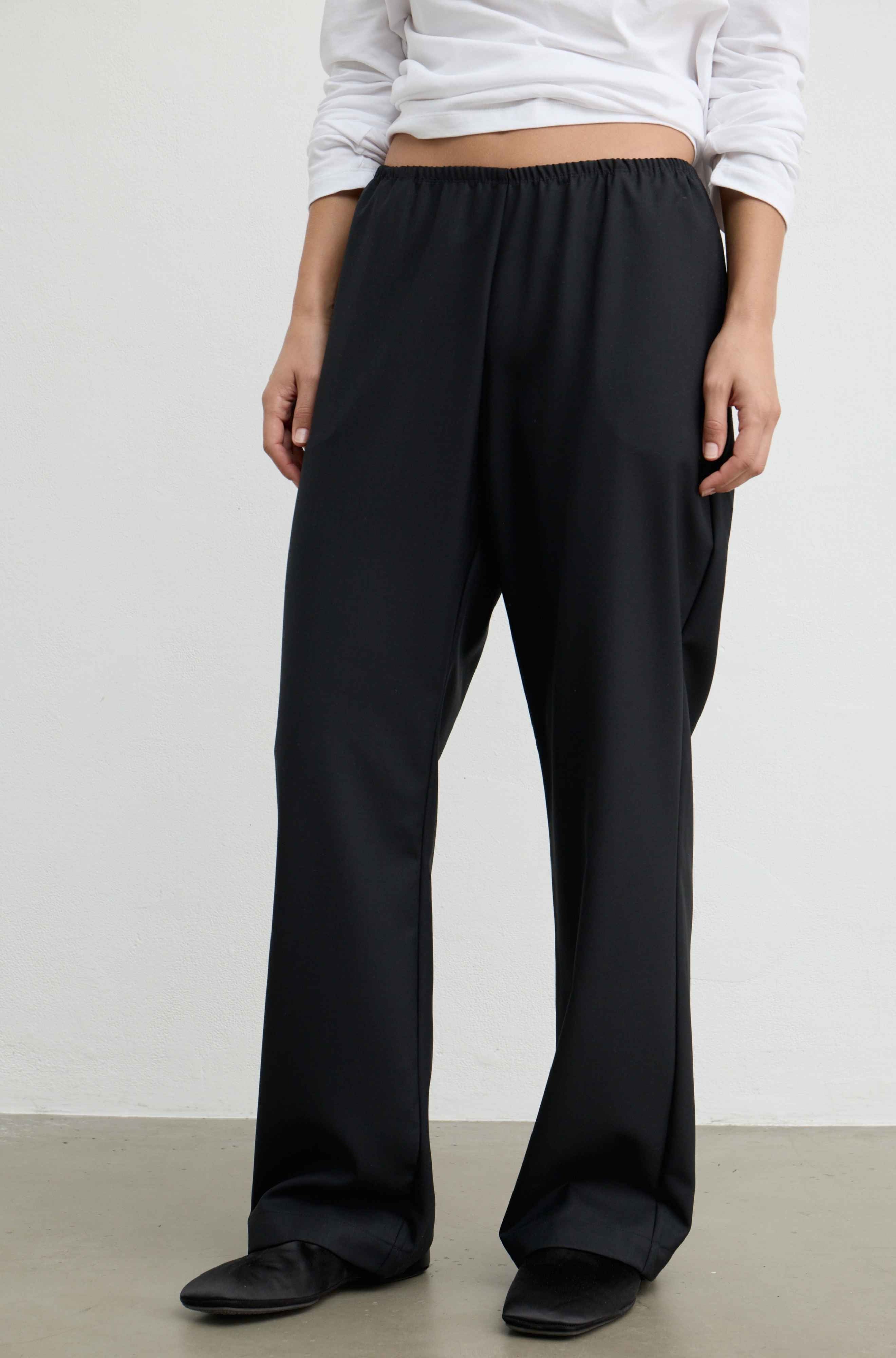 A person wearing loose black wide-leg trousers with an elastic waistband, paired with a white cropped long-sleeve top and black shoes, standing against a plain light gray wall.