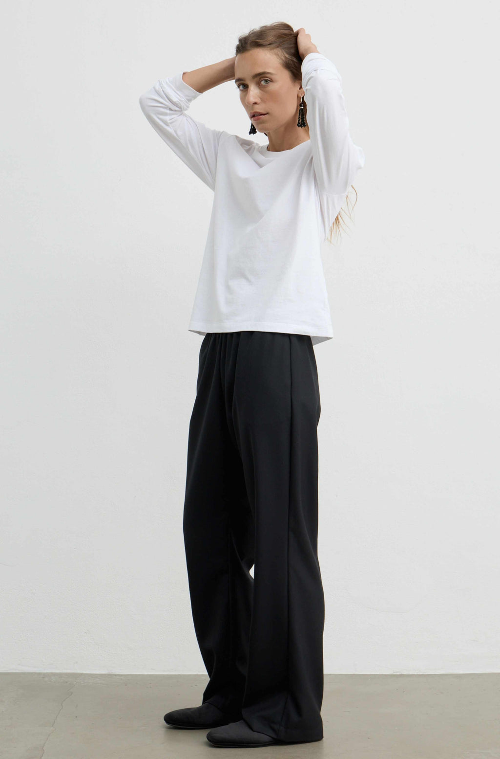 A woman modeling a white long-sleeve crewneck top and black wide-leg trousers against a plain white studio background.