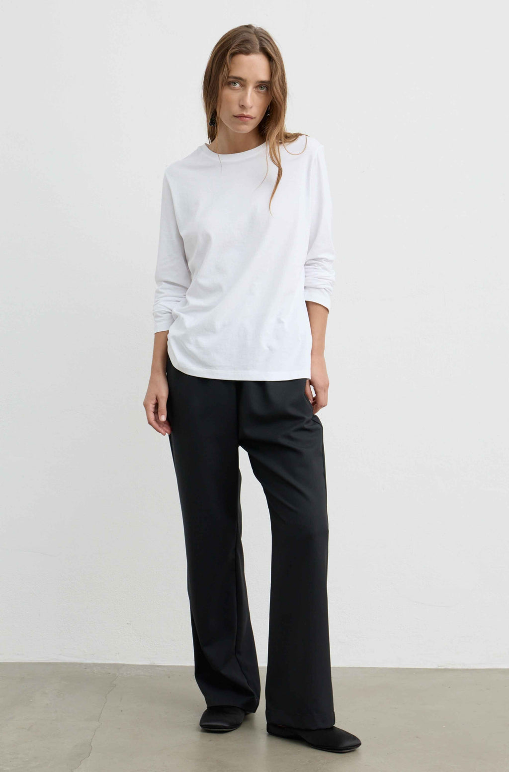 A woman modeling a white long-sleeve crewneck top and black wide-leg trousers against a minimalist white studio wall and concrete floor.