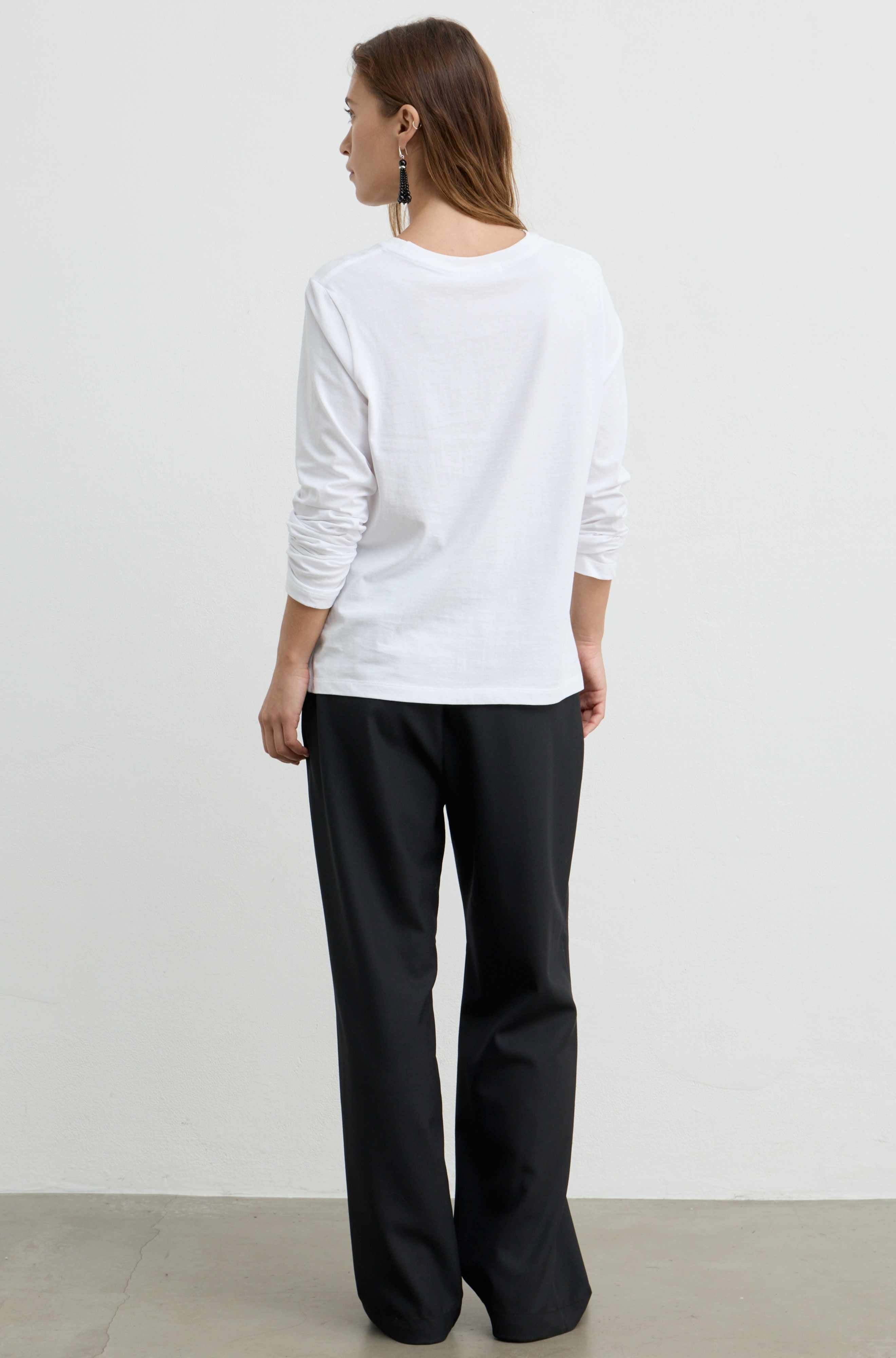 A woman is shown from the back wearing a white long-sleeve crewneck top and black wide-leg trousers, standing against a plain white wall.
