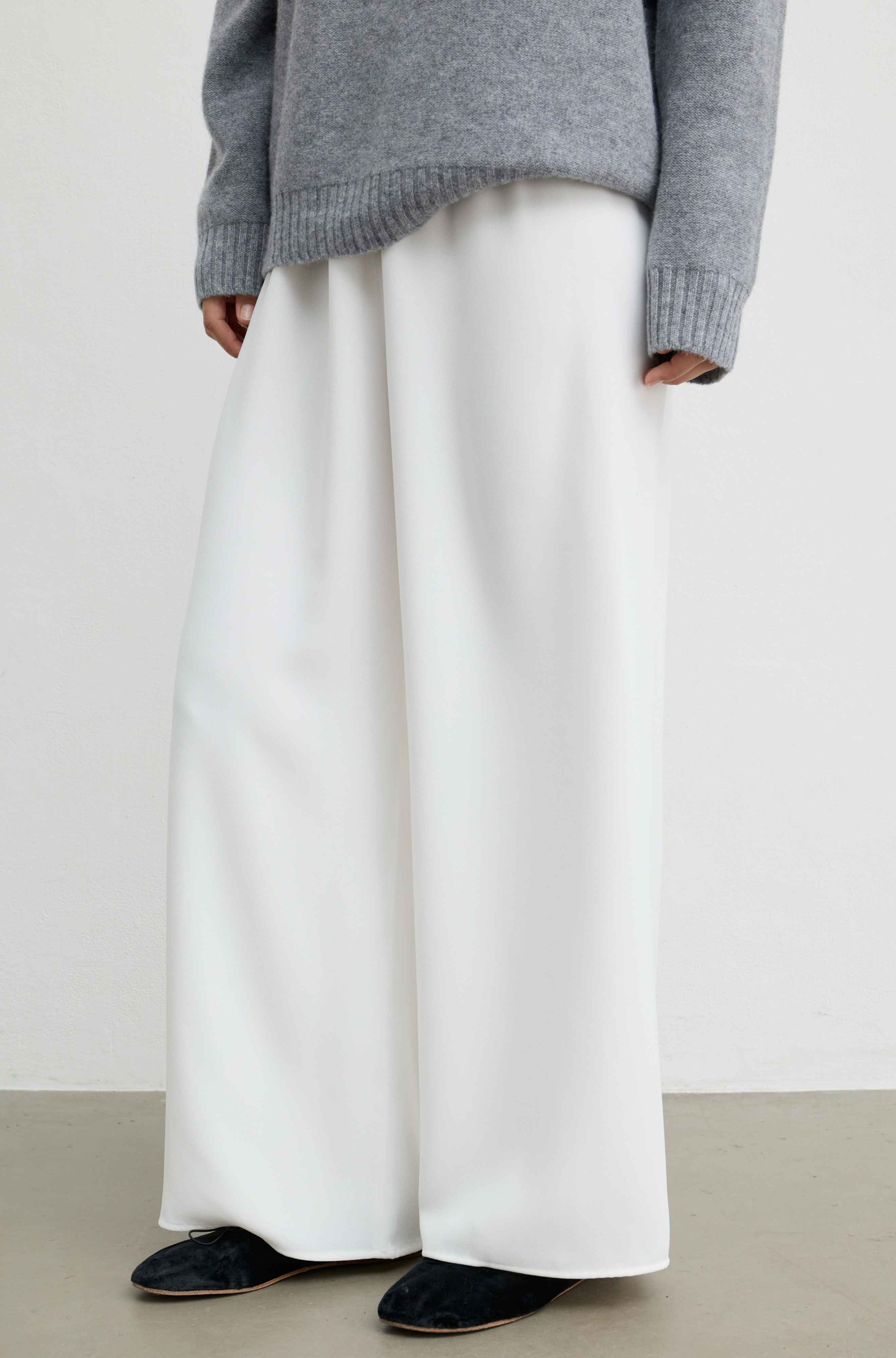 A person wearing wide-leg white trousers, a gray knit sweater, and black slippers against a plain light gray wall and concrete floor.