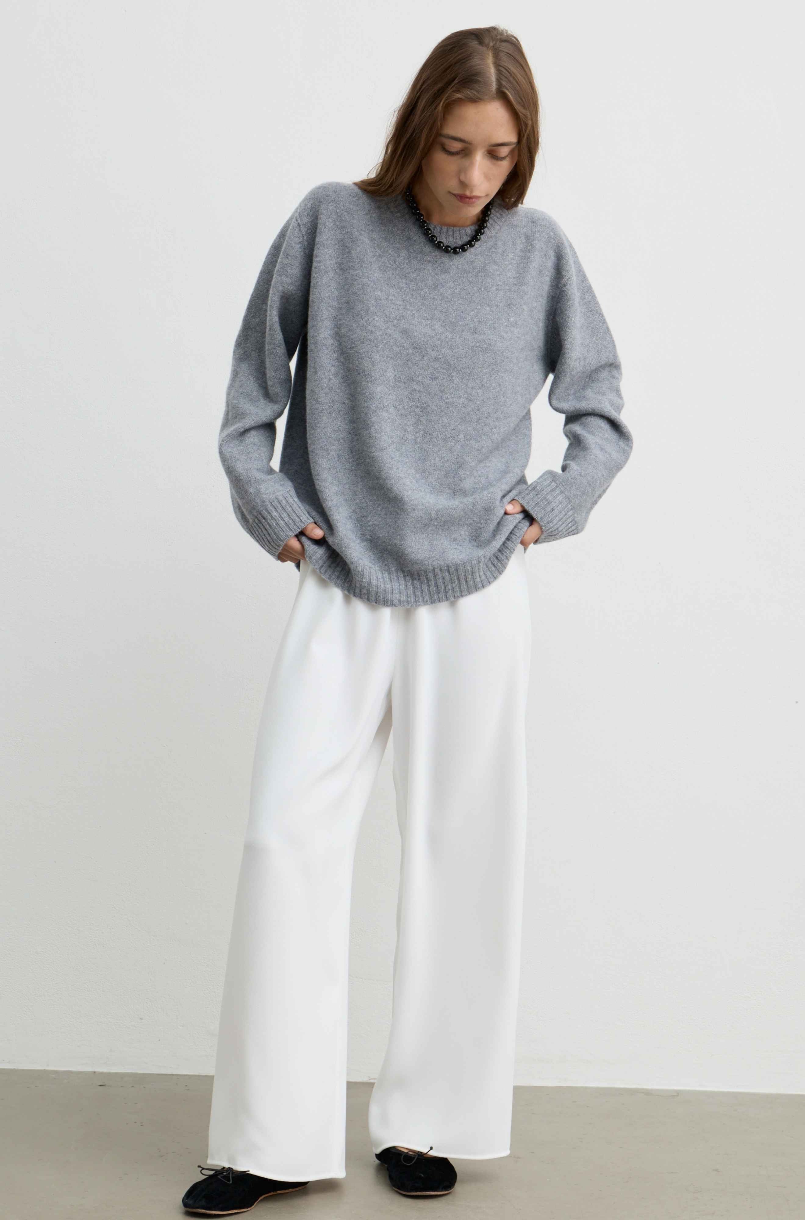 A woman wearing a loose-fit gray knit sweater, white wide-leg trousers, and black shoes, standing against a plain white studio wall.