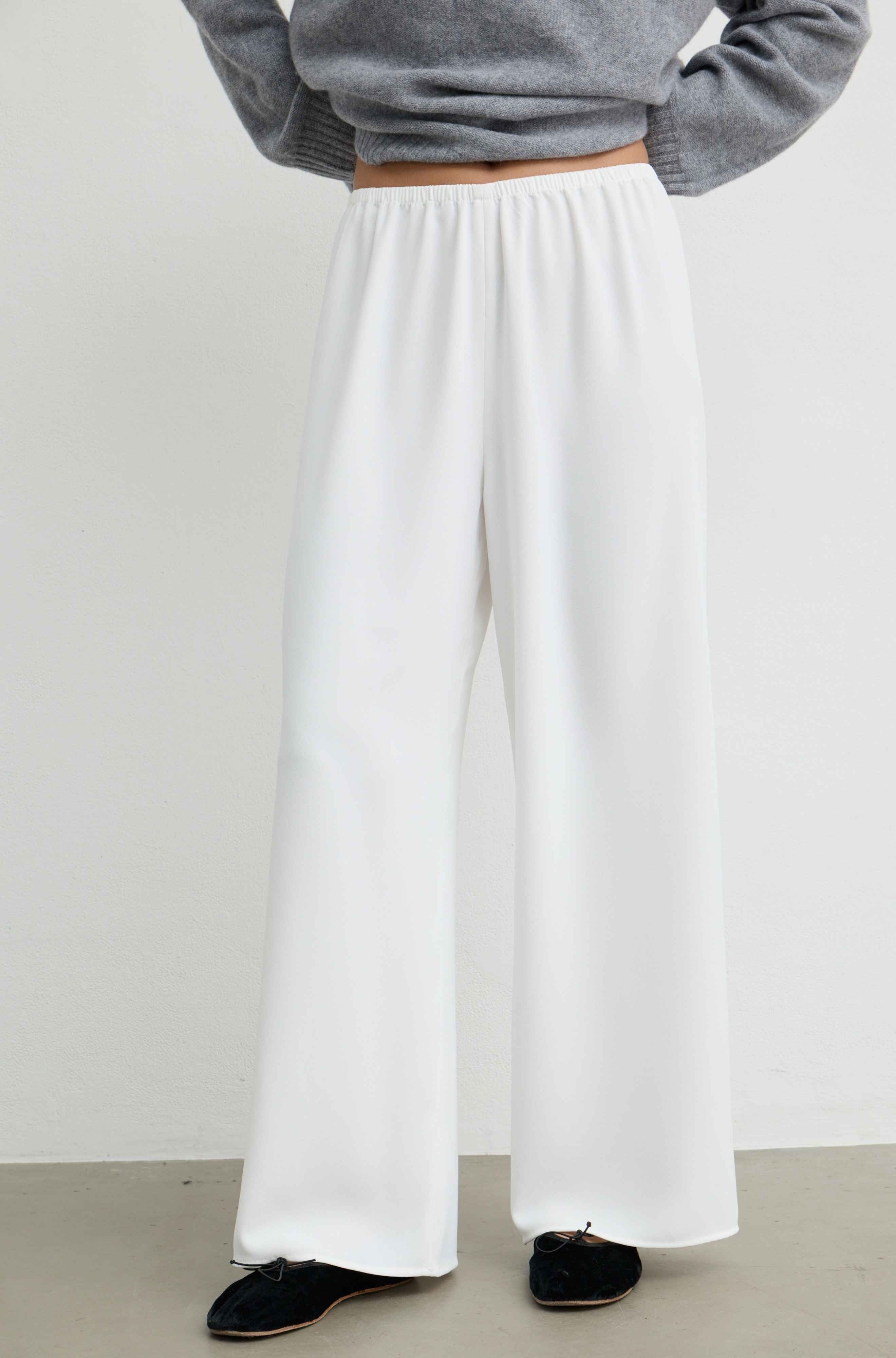 White wide-leg trousers worn by a model, paired with a gray long-sleeve top and black slip-on shoes, shown from the waist down against a plain light gray wall.