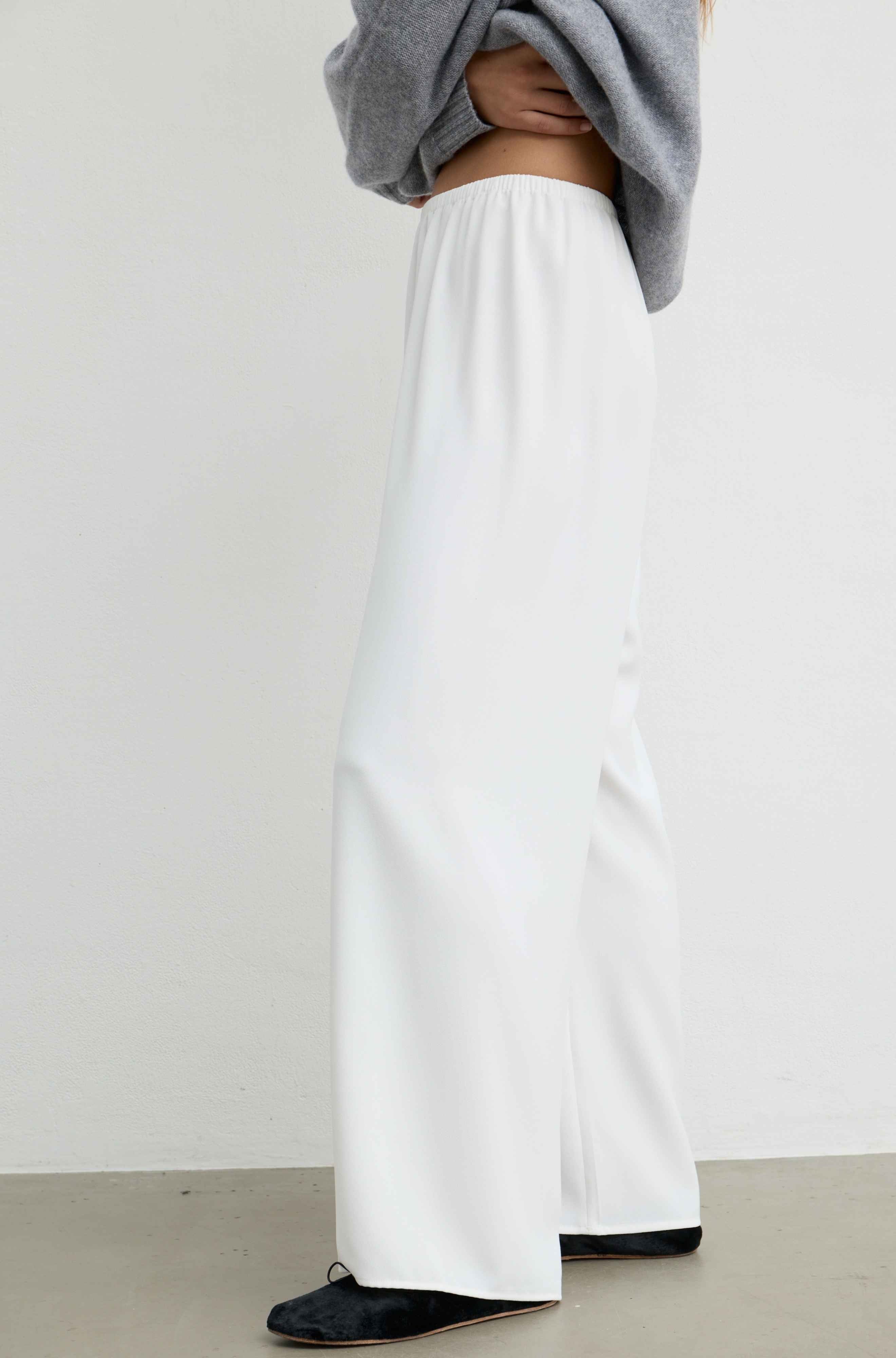 White wide-leg trousers worn by a model, styled with a gray cropped sweater and black flats, shown from the waist down against a plain light gray wall.