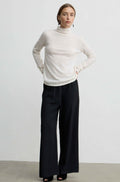 A woman modeling a cream turtleneck sweater and black wide-leg trousers against a plain light gray studio background.
