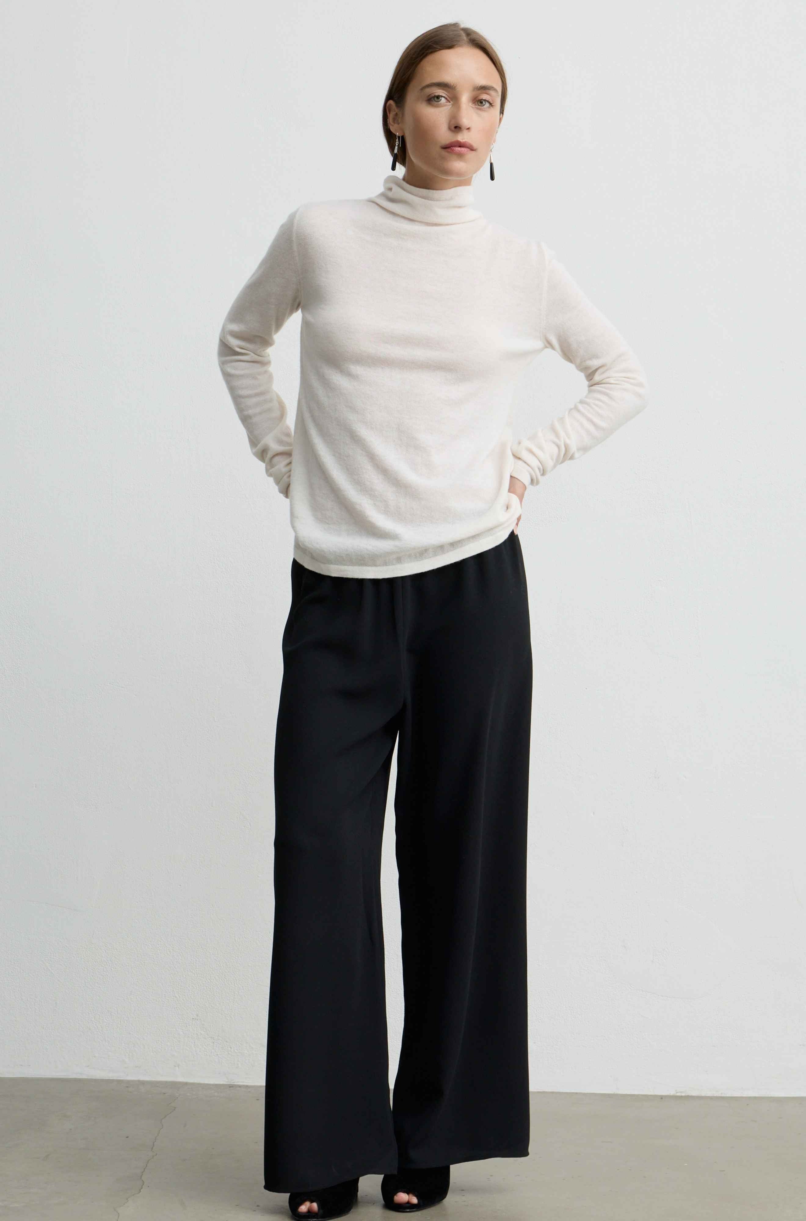 A woman modeling a cream turtleneck sweater and black wide-leg trousers against a plain light gray studio background.
