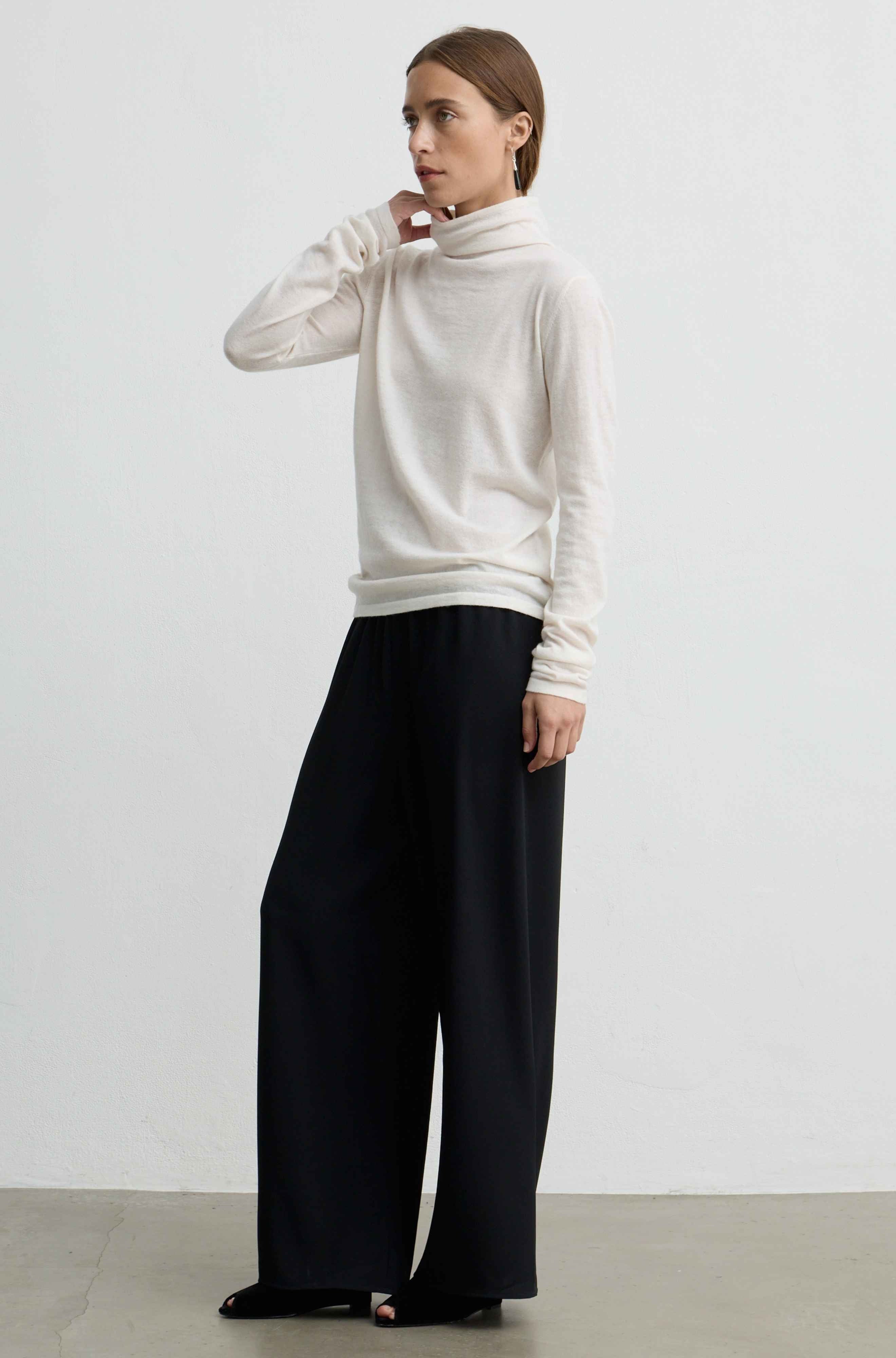 A woman modeling a cream-colored turtleneck sweater and black wide-leg trousers against a plain light gray studio background.