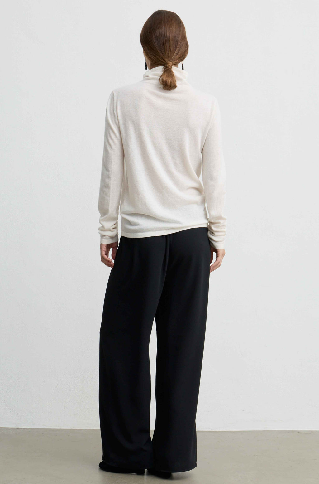 A woman viewed from behind wearing a cream-colored turtleneck sweater and black wide-leg trousers, standing against a plain white wall.