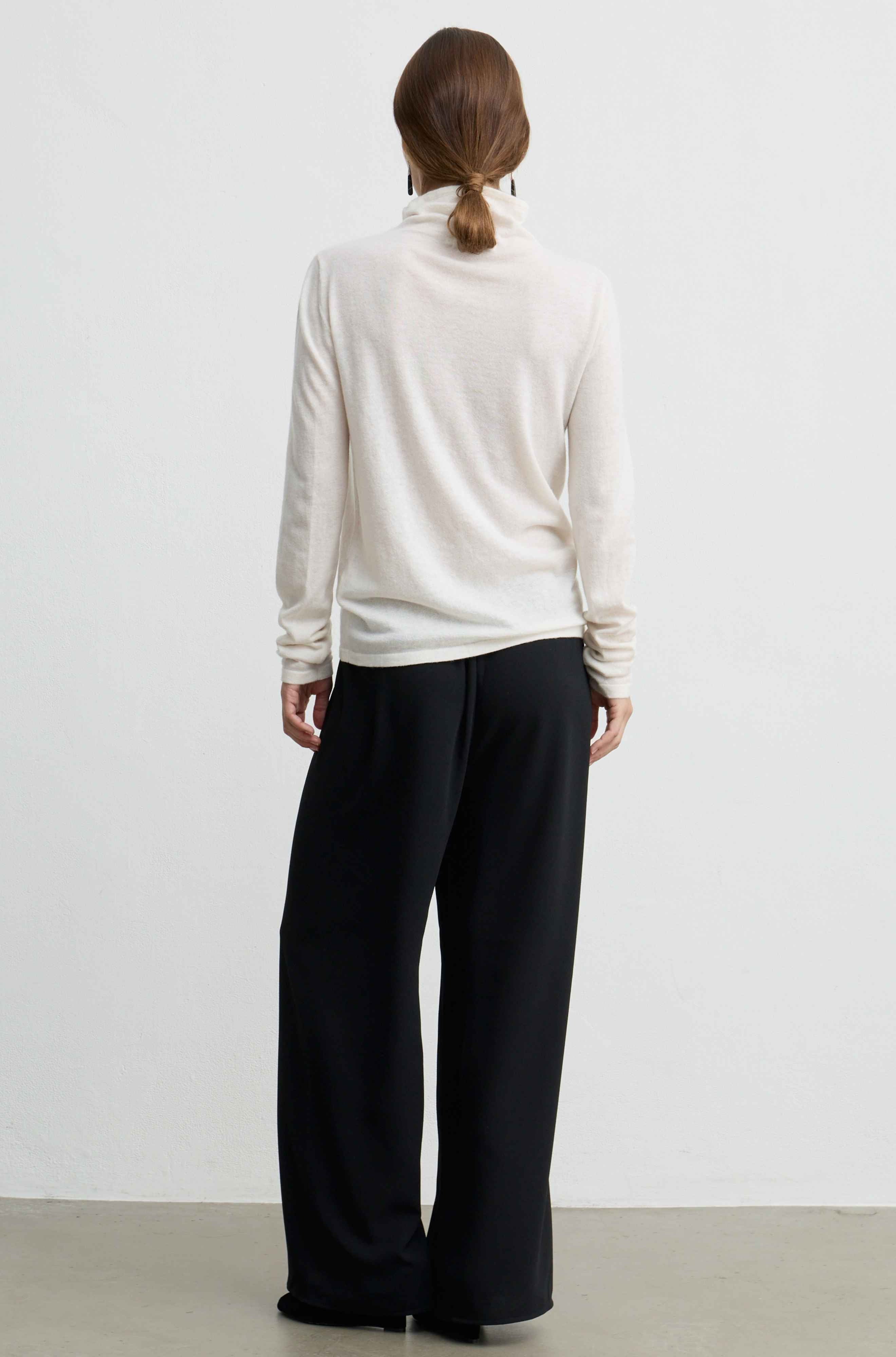 A woman viewed from behind wearing a cream-colored turtleneck sweater and black wide-leg trousers, standing against a plain white wall.