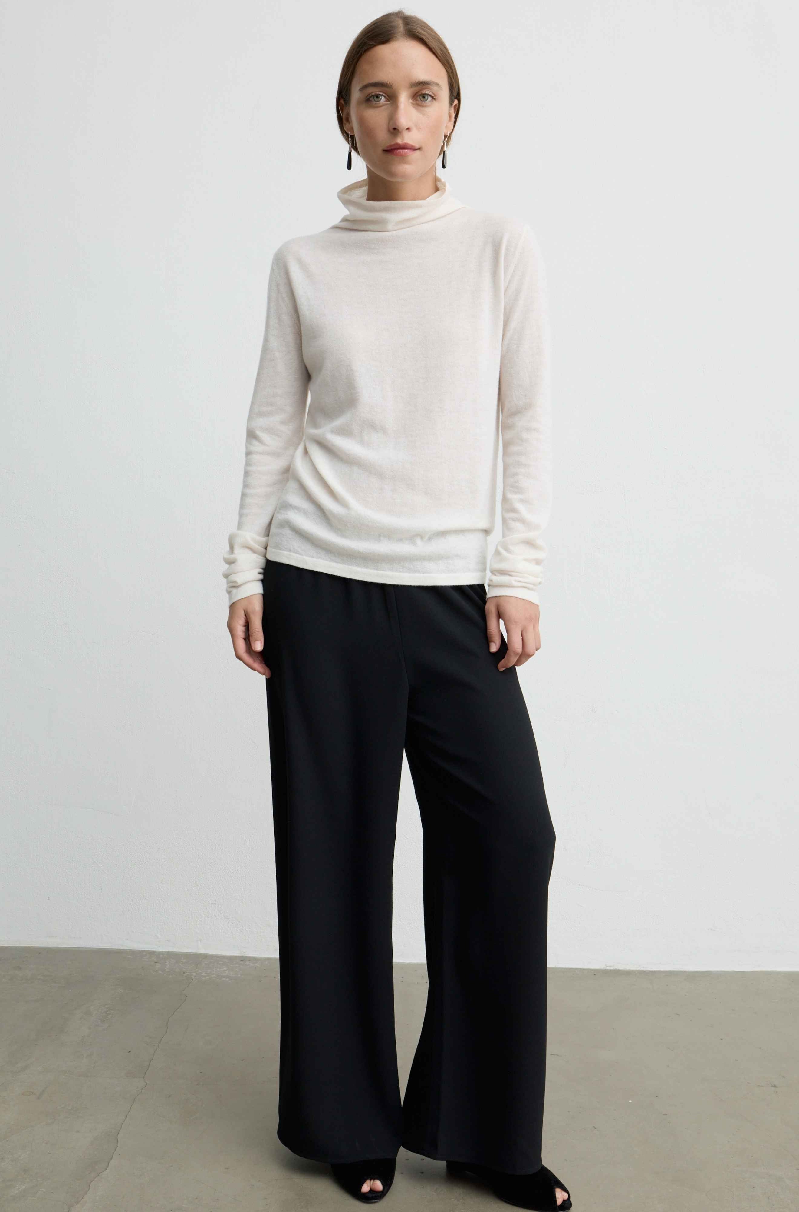 A woman modeling a cream-colored long-sleeve turtleneck sweater paired with black wide-leg trousers, standing against a minimalist light gray studio wall.
