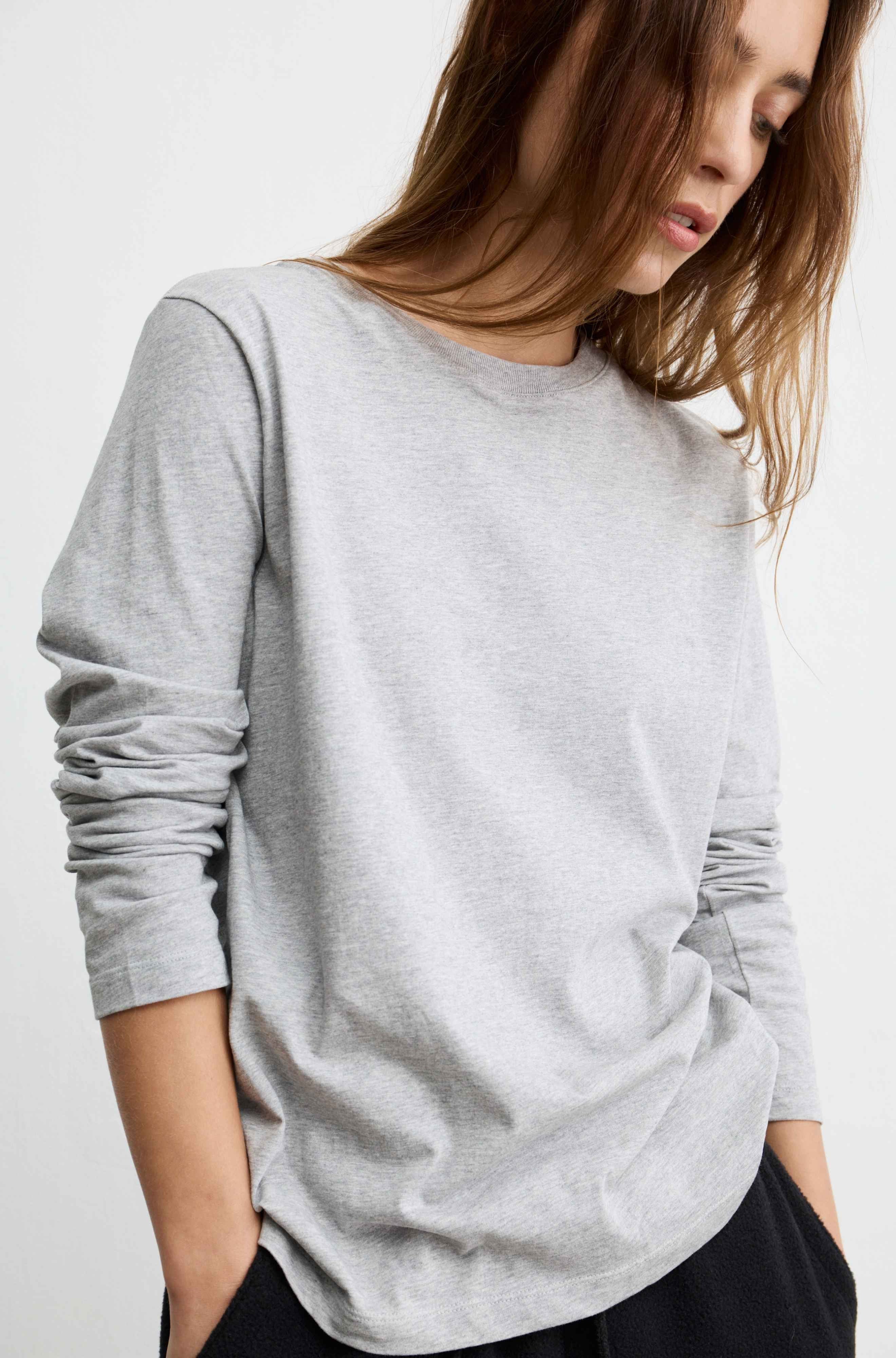 A woman wearing a light gray heather crewneck long-sleeve top with ruched sleeves, paired with black bottoms, against a plain white background.