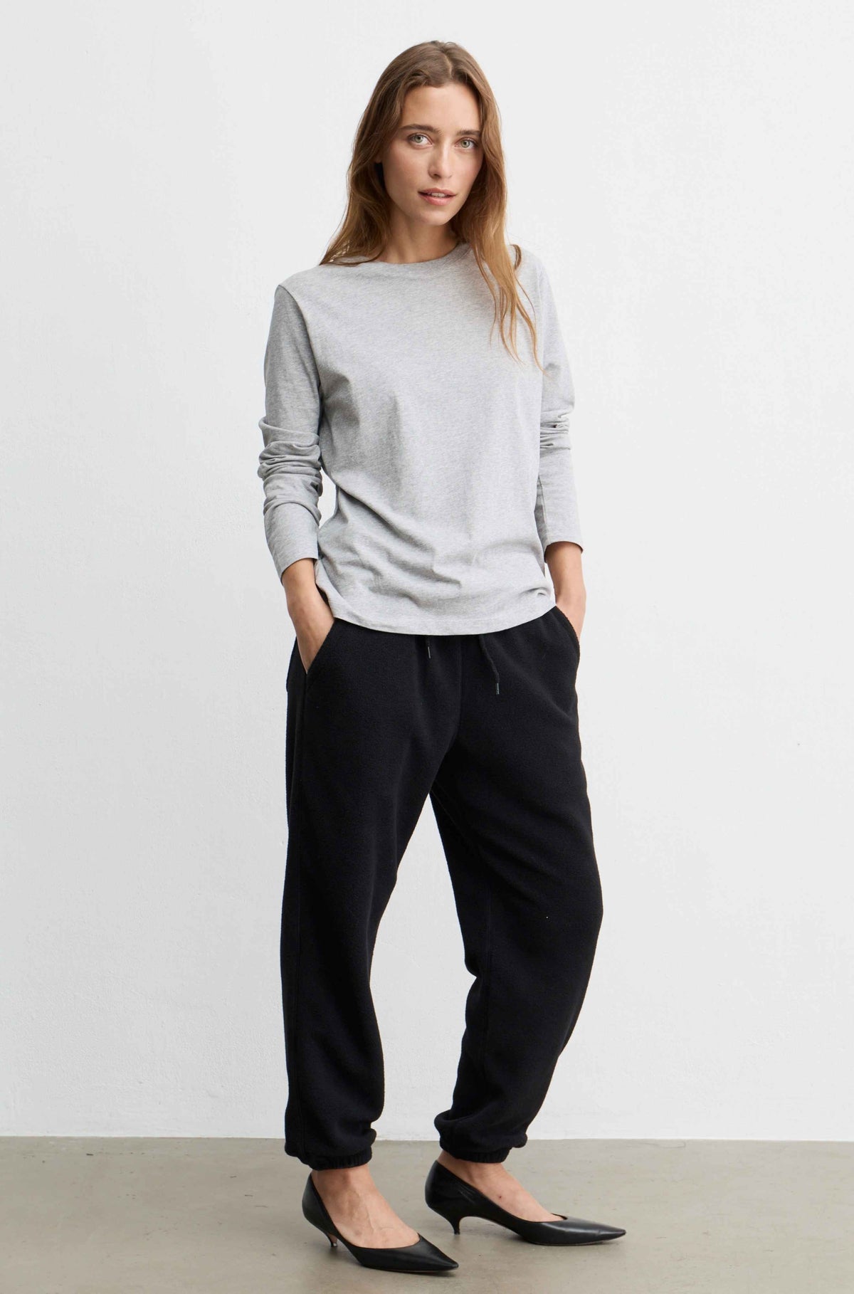 A woman modeling a light gray long-sleeve crewneck top and black jogger pants with black pointed-toe heels, standing against a plain white studio wall.