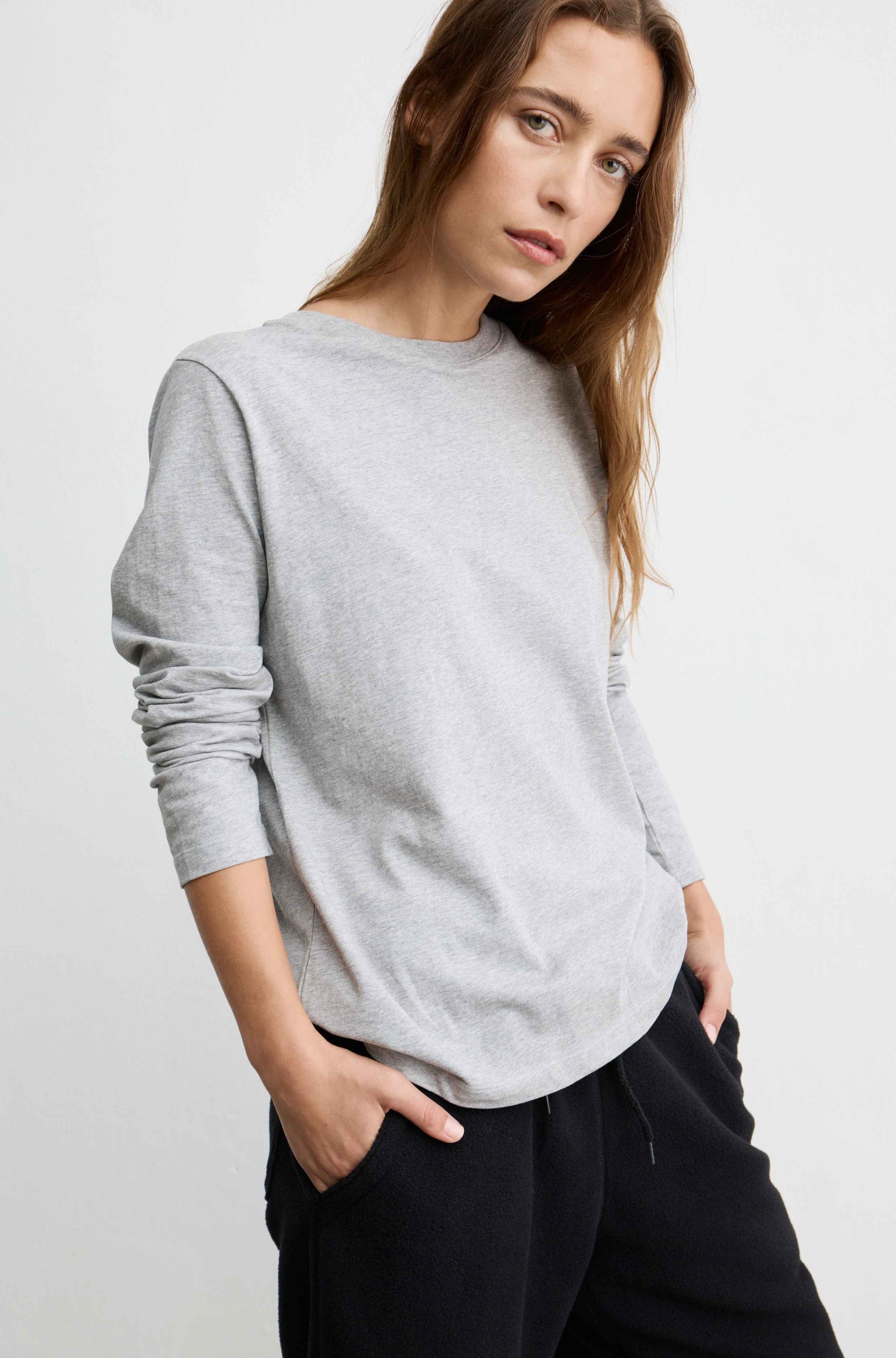 A woman wearing a light gray long-sleeve crewneck sweatshirt and black sweatpants, posing against a light gray studio background.